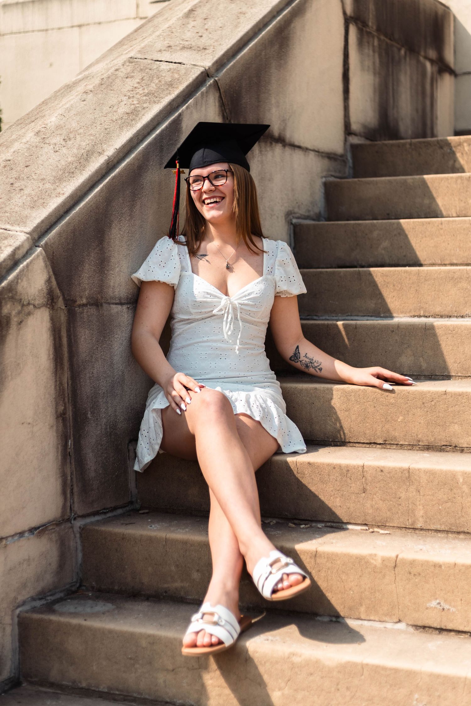 Boyertown Senior graduate in white dress and mortarboard cap sitting on stone steps laughing in sunlight.