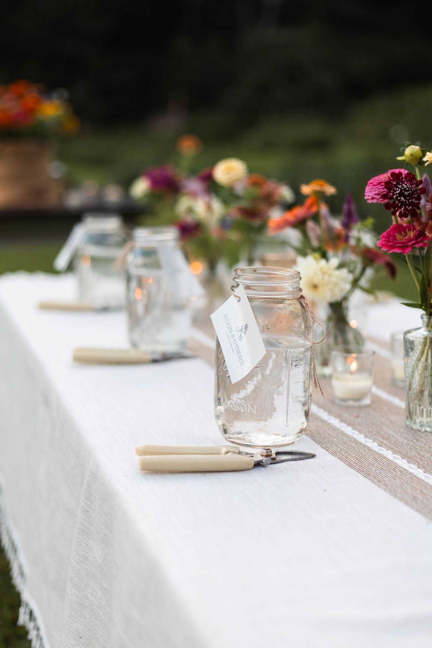 Rustic outdoor dining table set with mason jar glasses, taper candles, and colorful wildflower arrangements on white linen.
