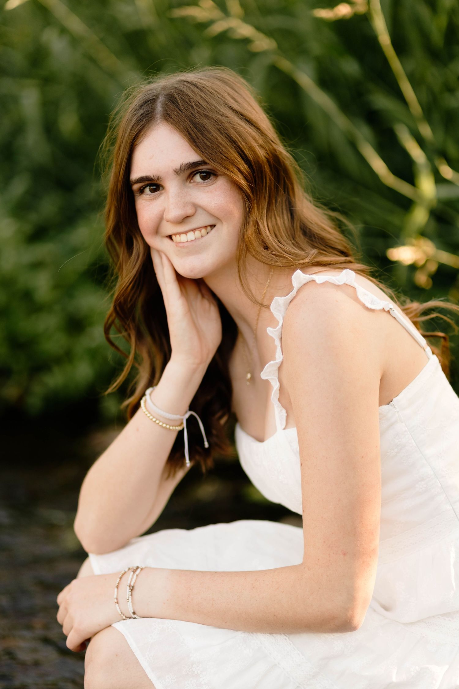 Smiling Boyertown High School Senior in white dress sitting near greenery, resting cheek on hand, wearing pearl bracelet.