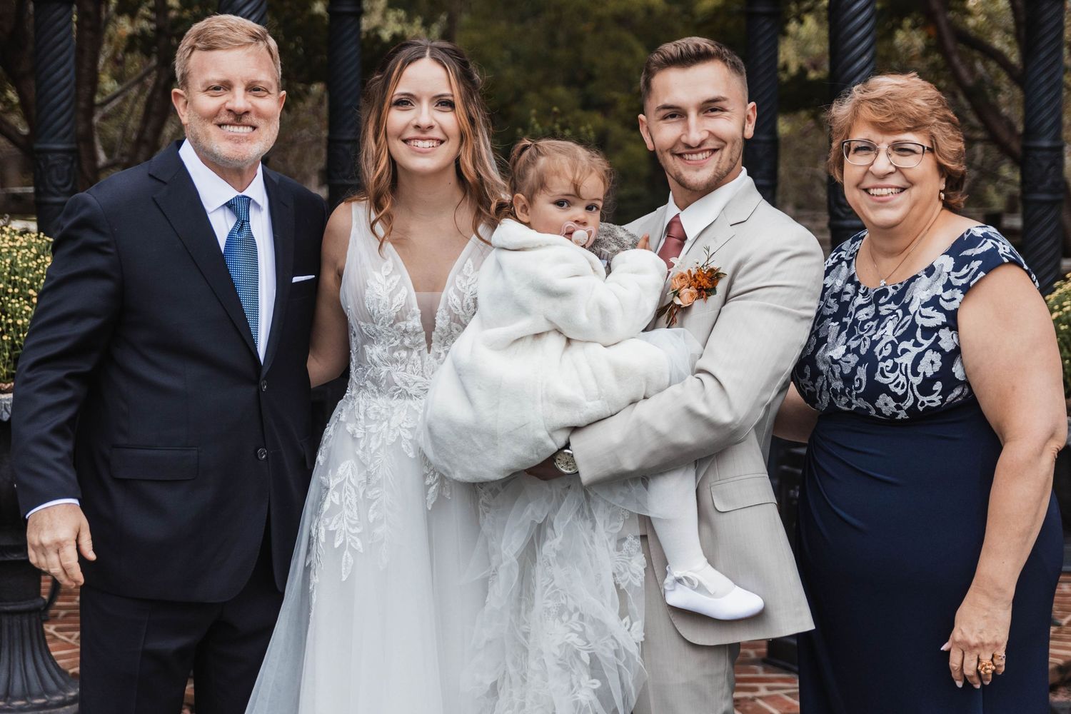 Newlyweds pose with small child and family members in formal attire at an outdoor wedding venue.