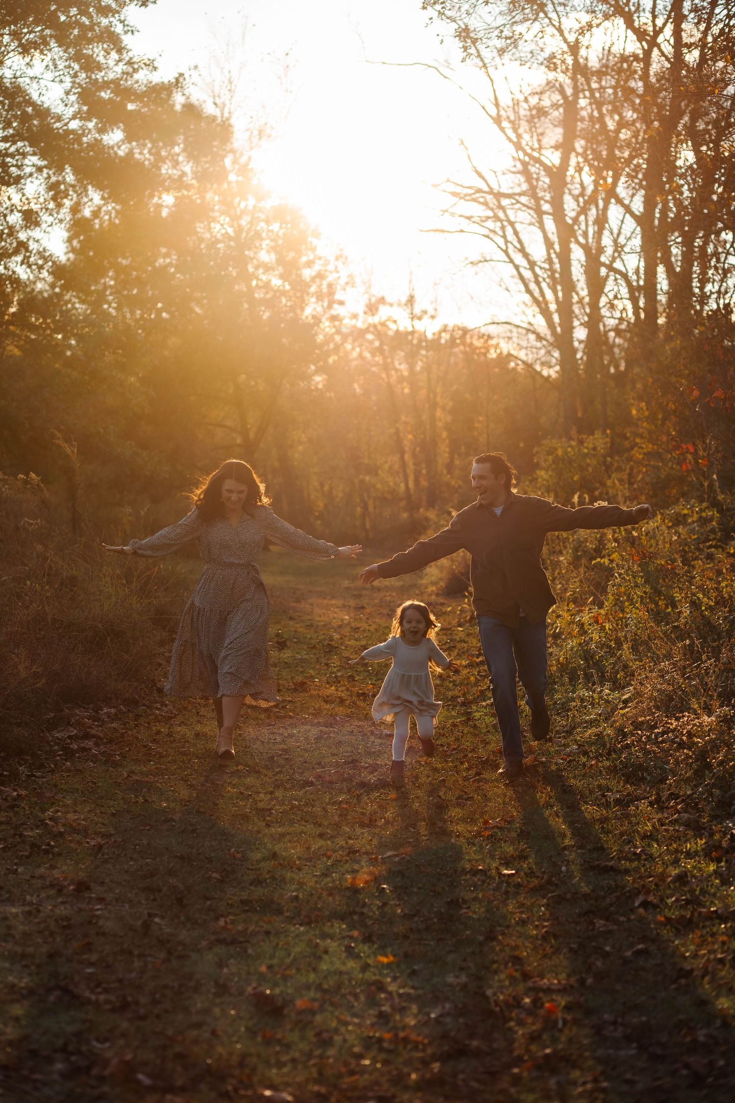 Family running on a sunlit autumn forest path at golden hour, parents and child playing outdoors.