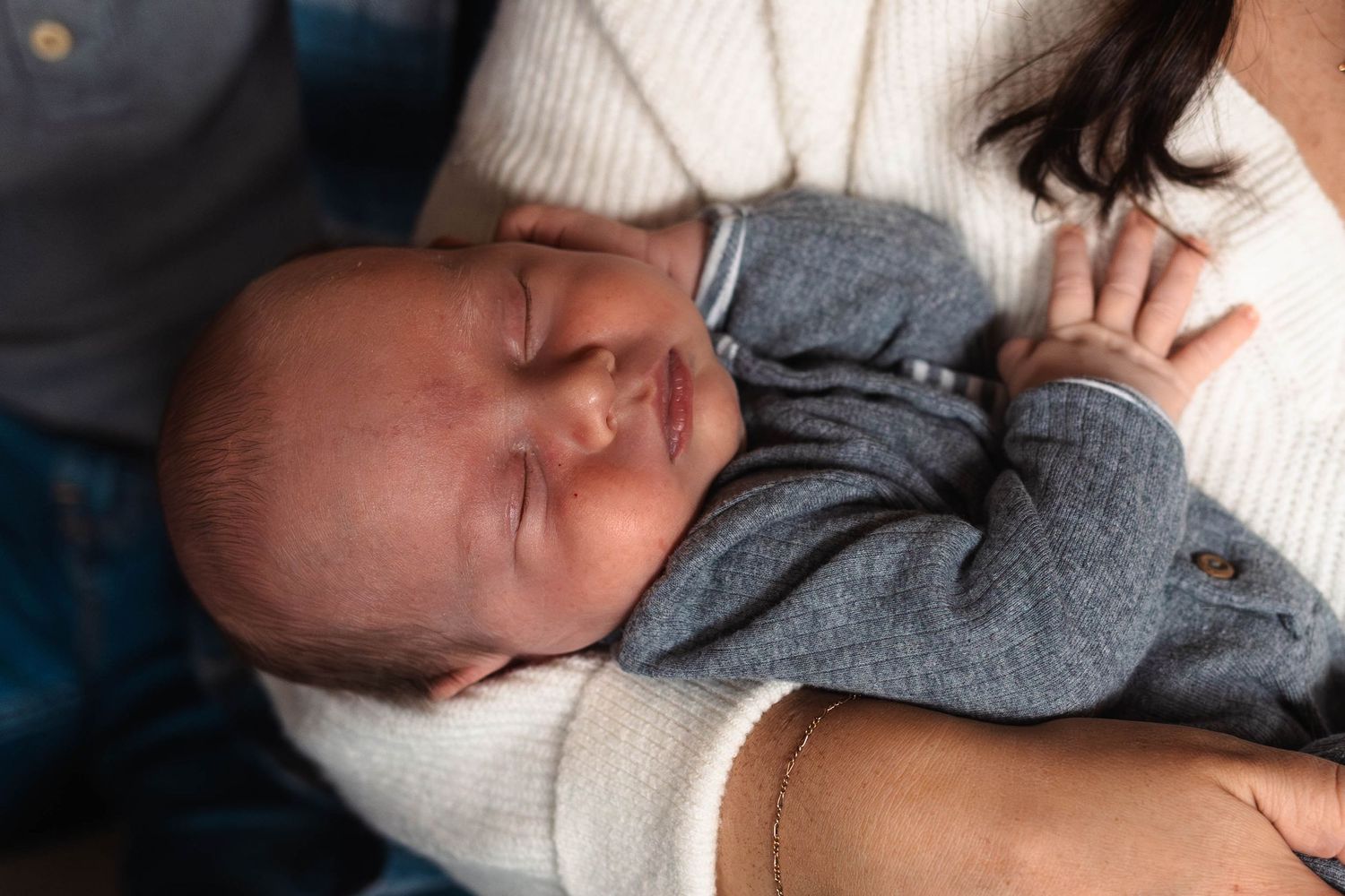Sleeping newborn baby in a gray outfit cradled in mothers arms wearing a white sweater.