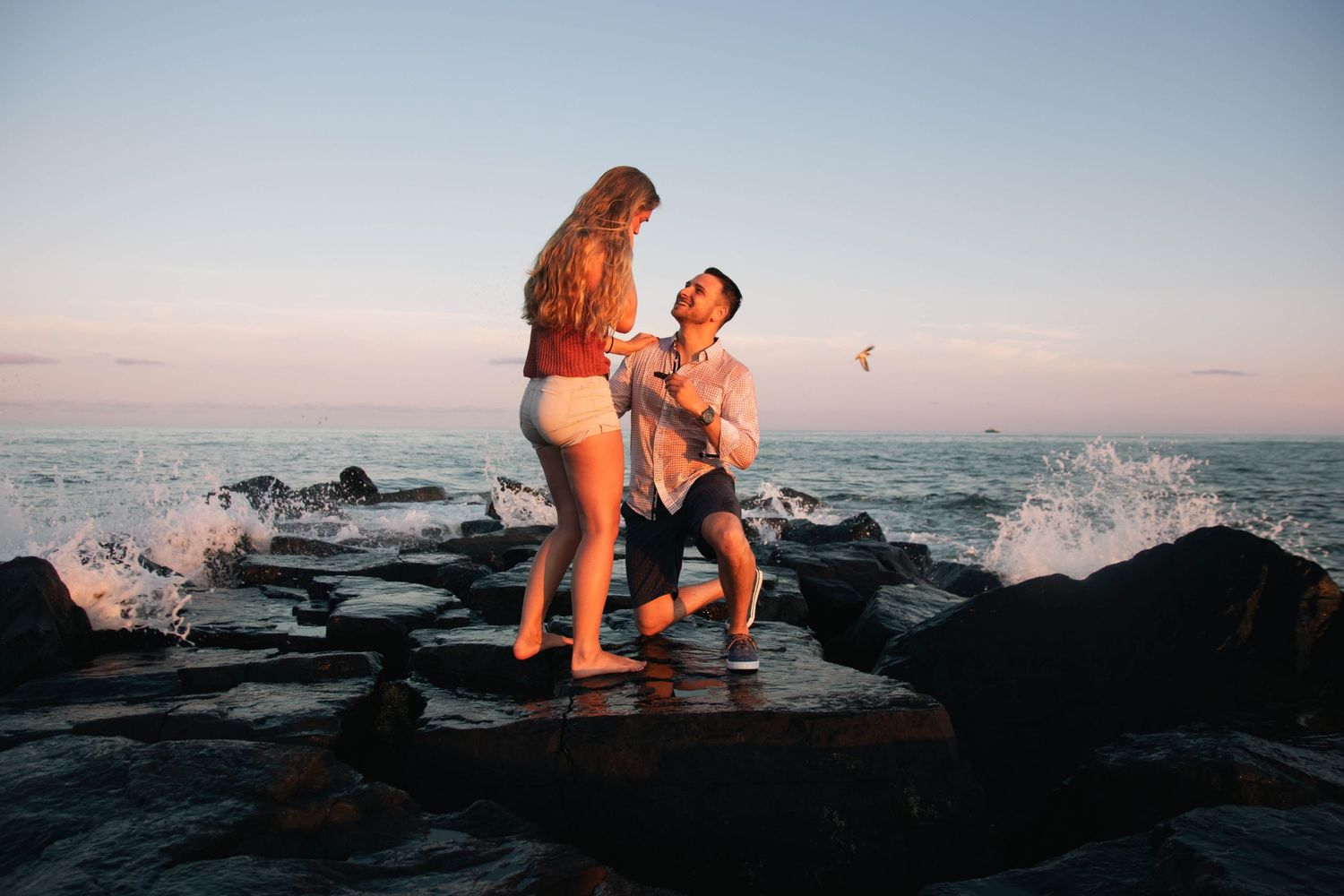 Romantic beach proposal at sunset with man on one knee proposing to woman on rocky shoreline with ocean waves.