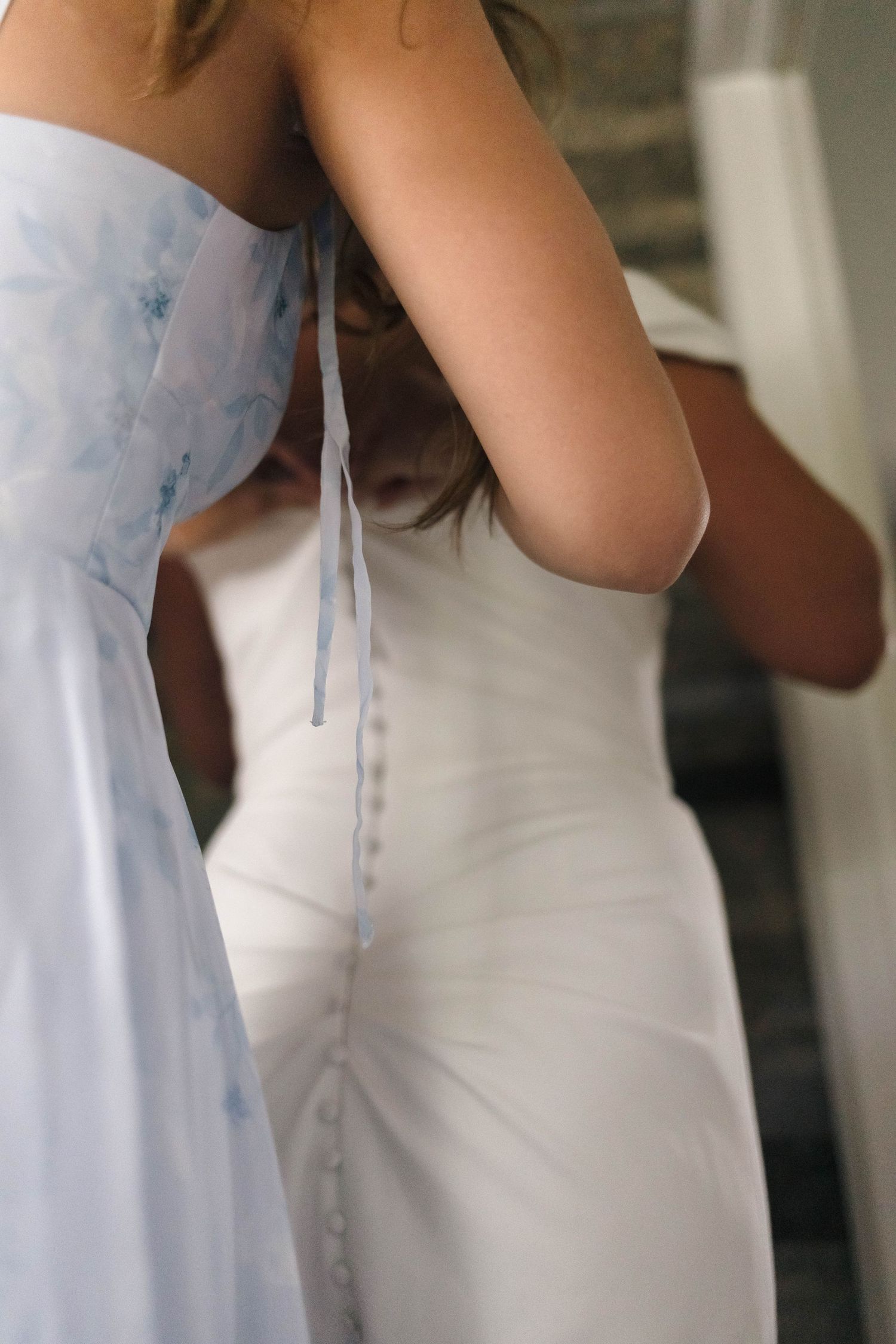 Close-up of bride in white button-back wedding dress being tied by bridesmaid in blue floral gown.