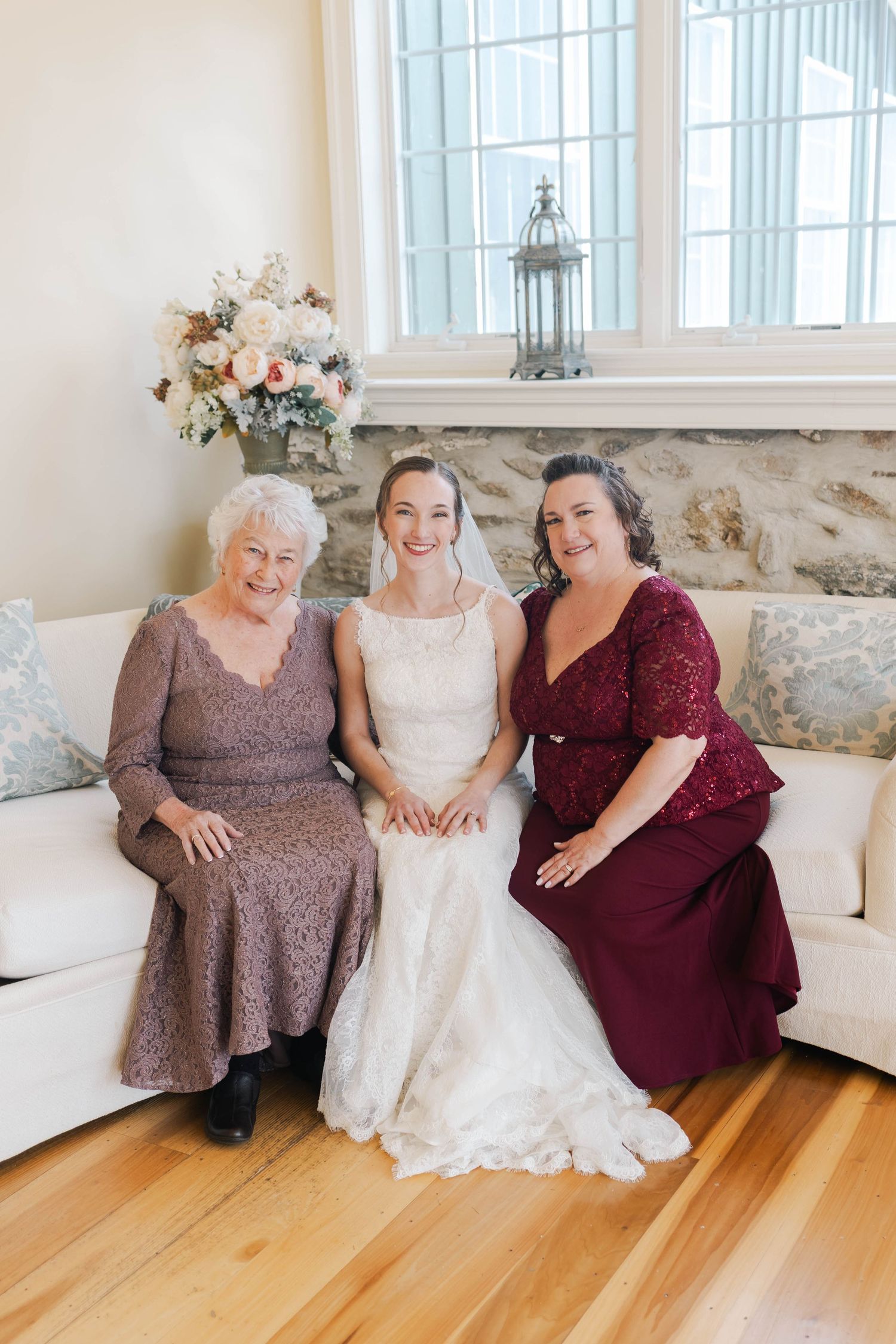 Bride in white lace gown sits smiling with her mother and grandmother on white sofa indoors.