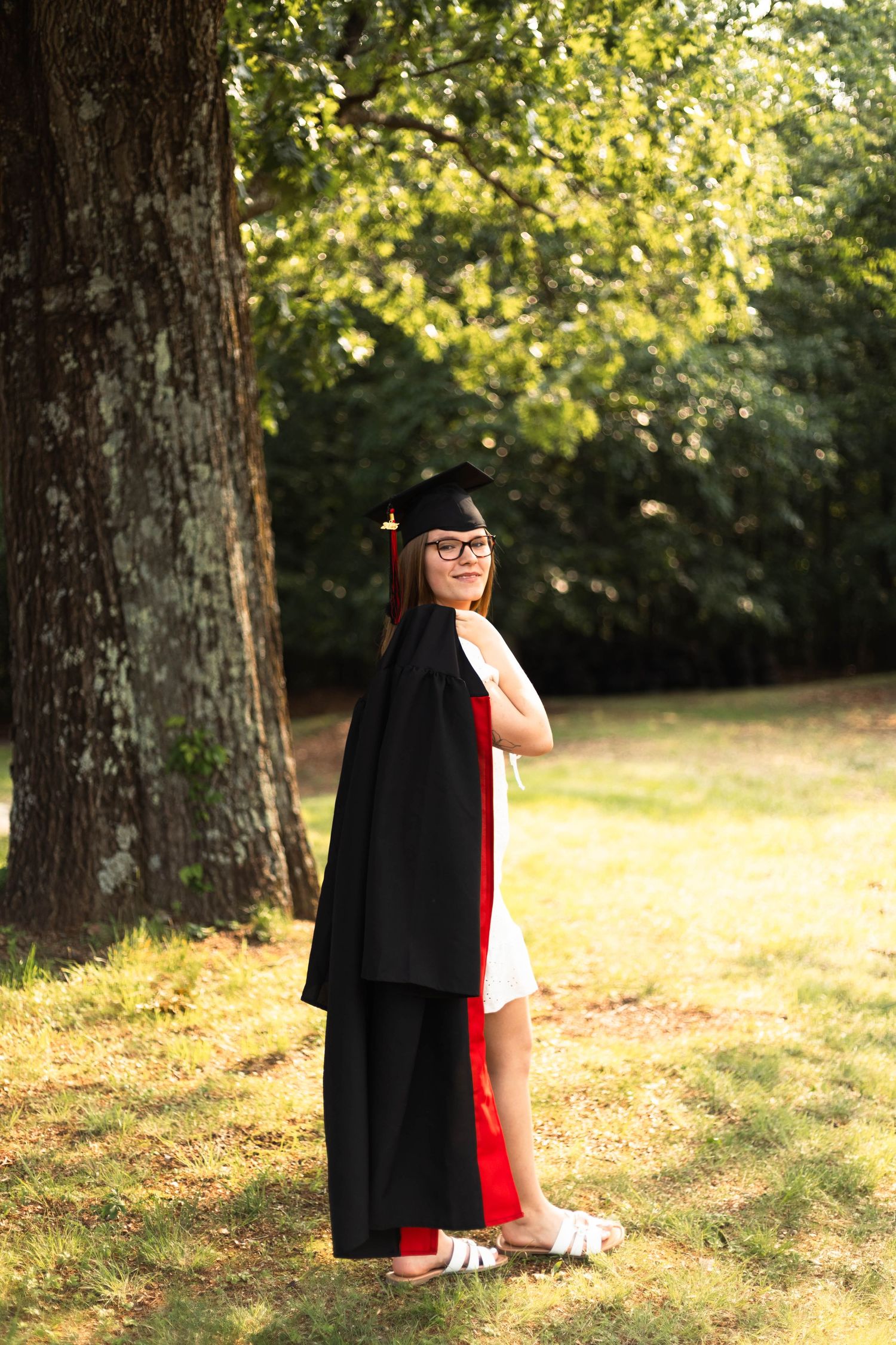 Boyertown graduate in black cap and gown with red sash standing outdoors near a large tree in sunlit park.