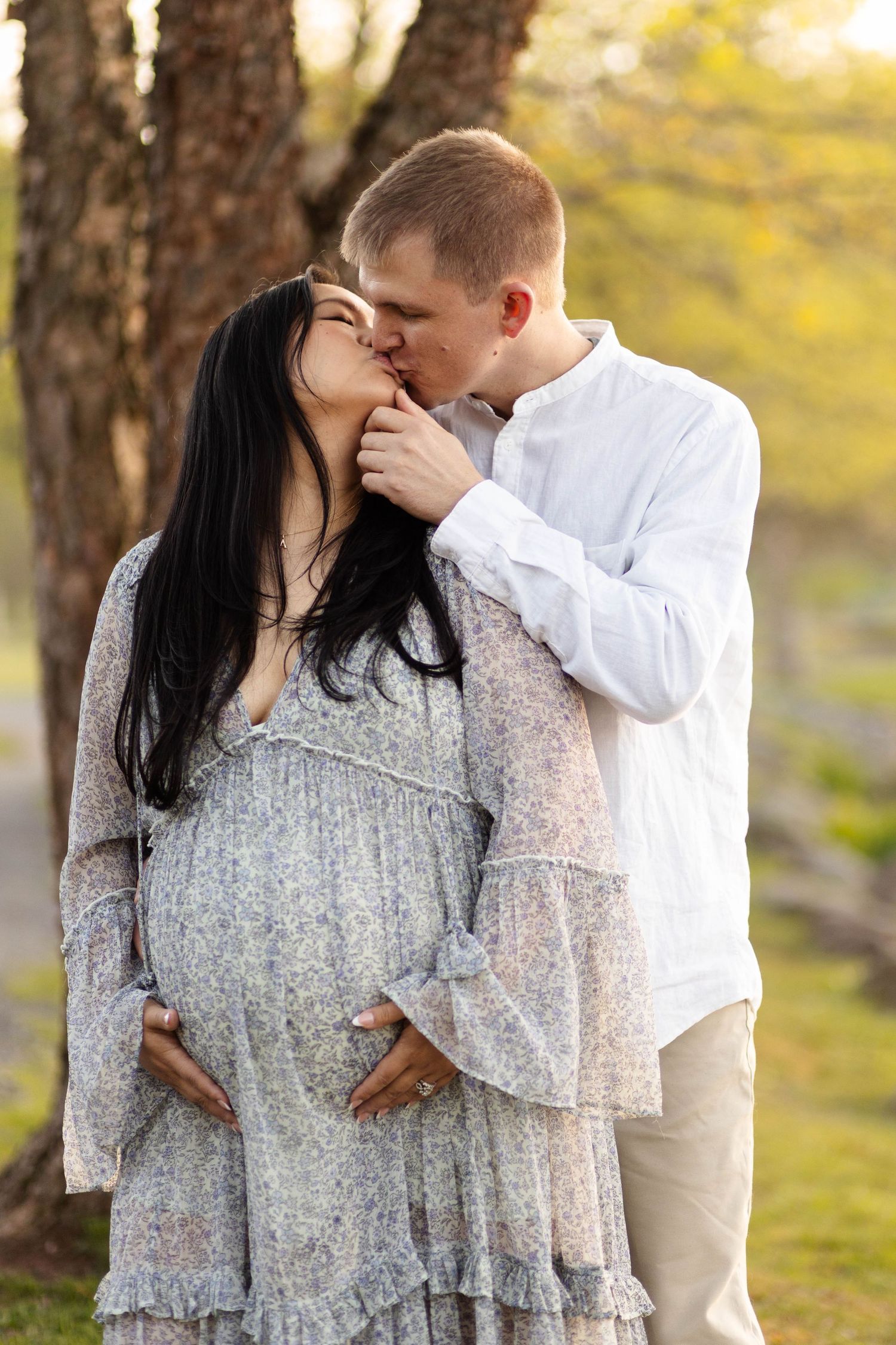 Pregnant woman in floral dress kissing partner outdoors during golden hour maternity photoshoot at Green Lane Park..