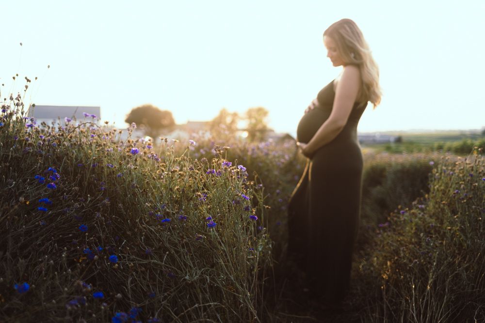 Pregnant woman in black dress standing in wildflower field during golden sunset light.