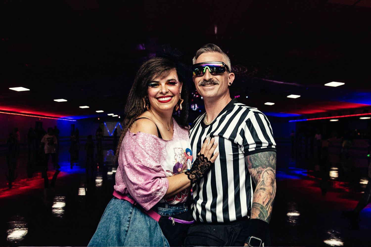 Smiling couple poses together at a roller skating rink wearing retro-inspired outfits under colorful lights.