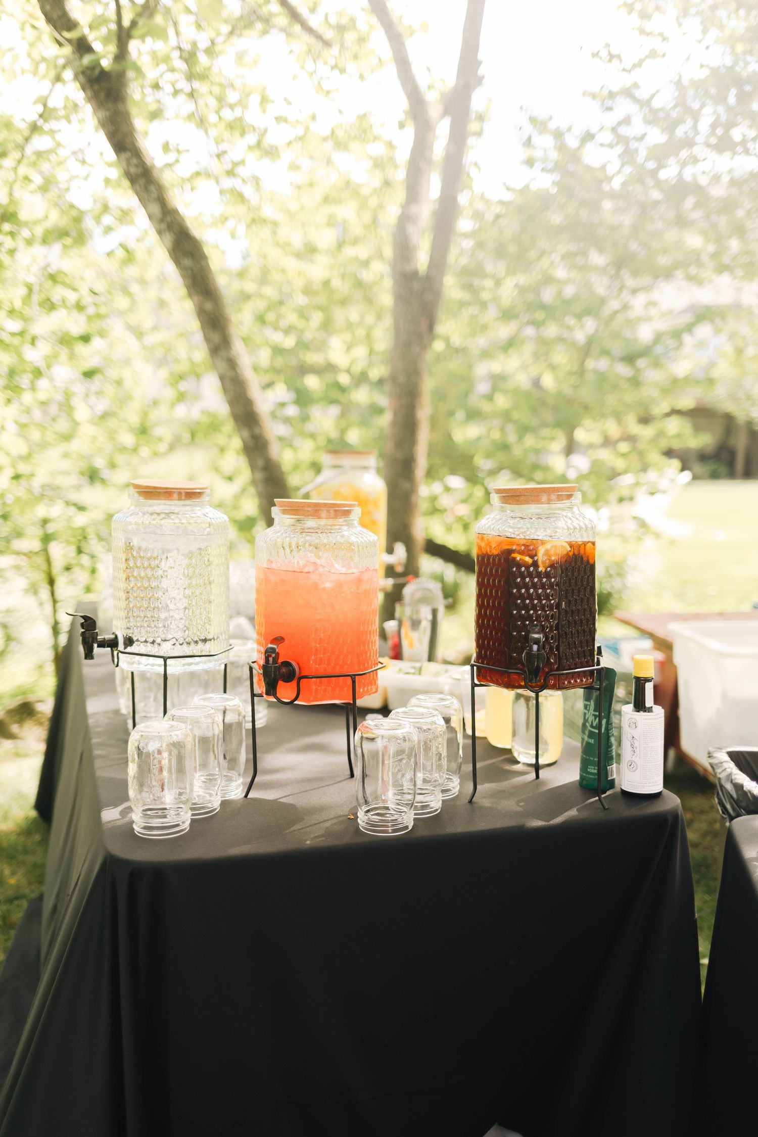 Outdoor wedding beverage station with three glass dispensers of pink and amber drinks on black table.