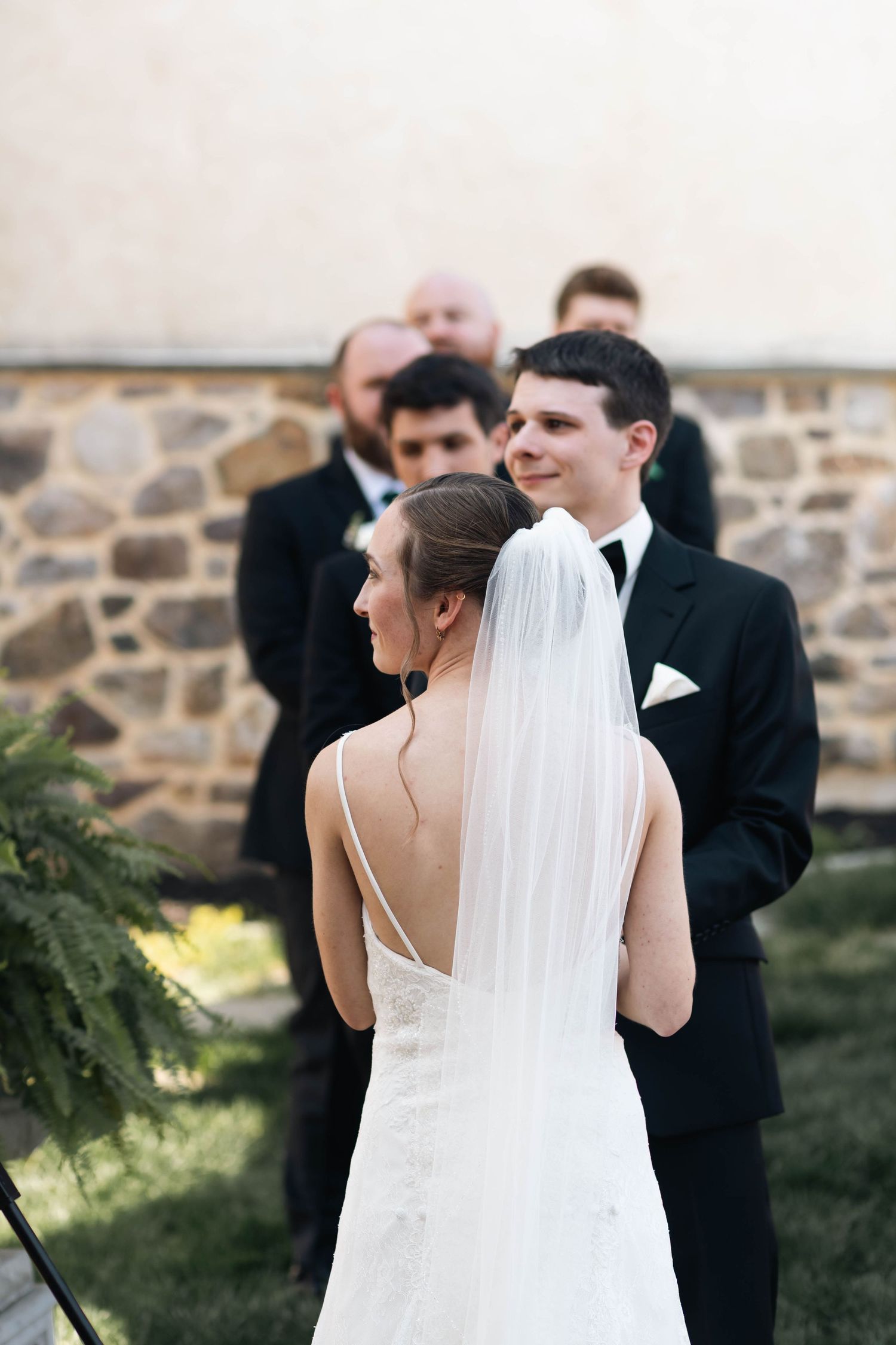 Groom smiles at bride standing at outdoor wedding altar with groomsmen and stone wall backdrop.