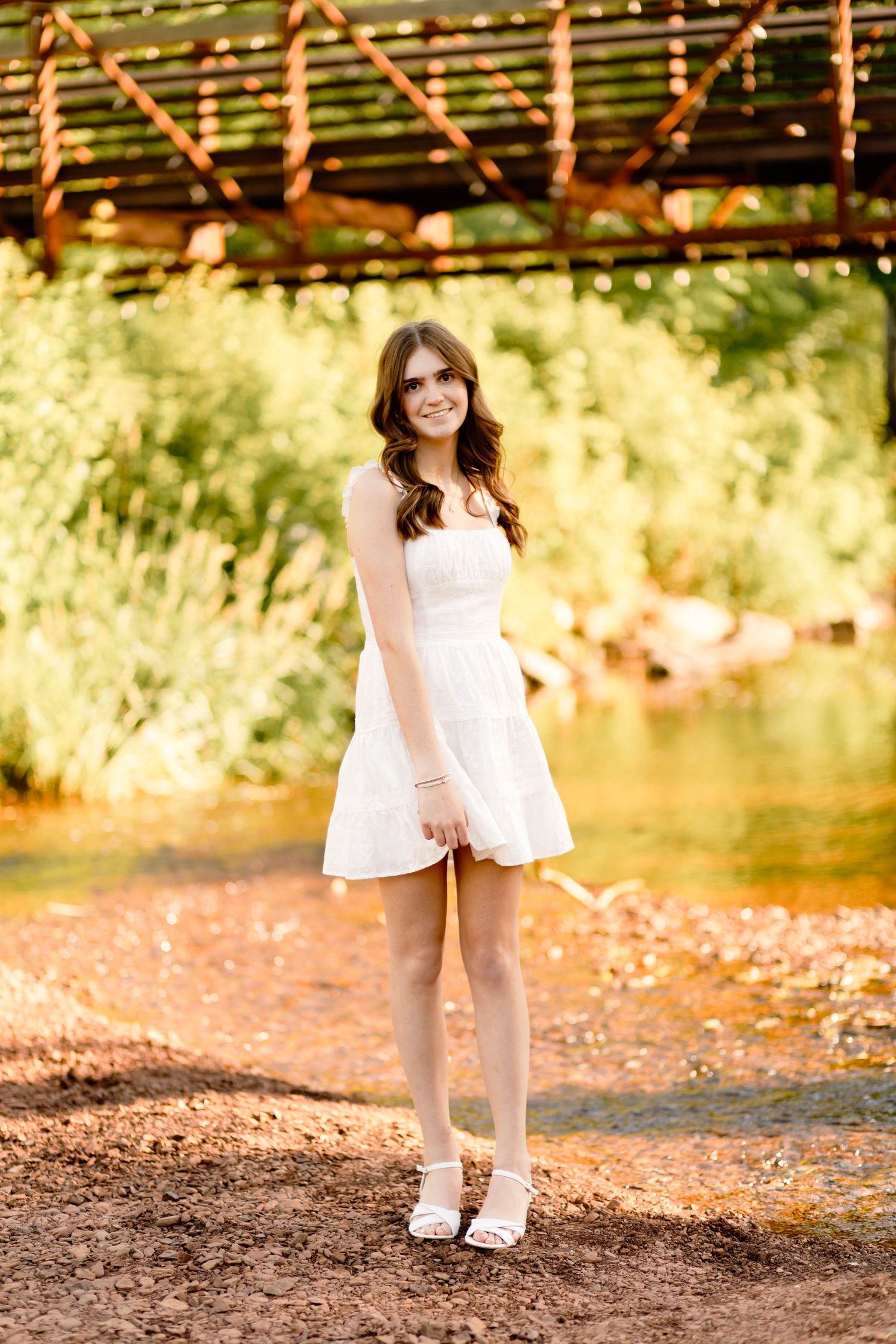 Boyertown Senior  in white sundress standing near a creek under a rustic bridge in golden sunlight.