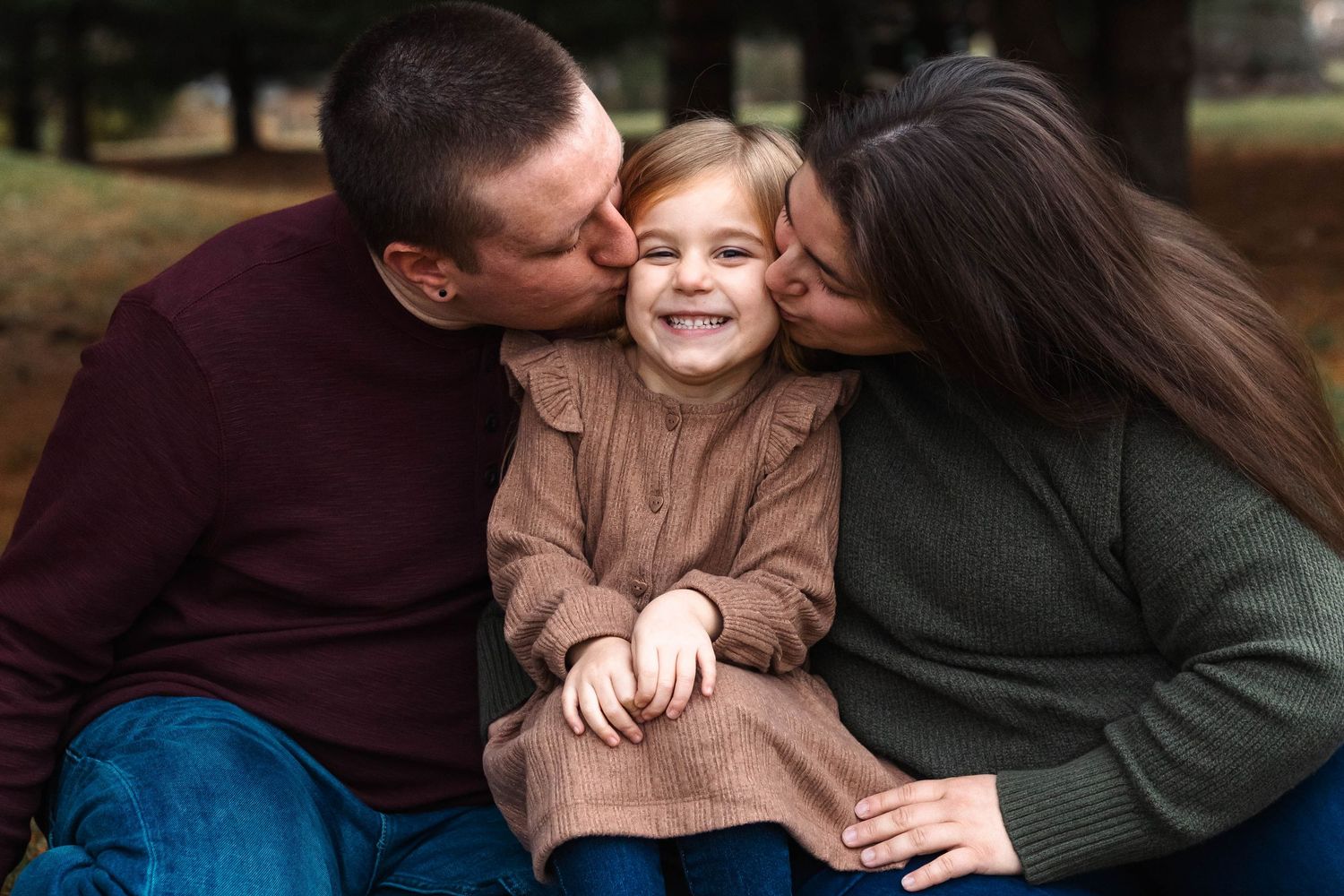 Happy toddler girl laughs as both parents kiss her cheeks simultaneously in a cozy outdoor portrait in Pottstown, PA.