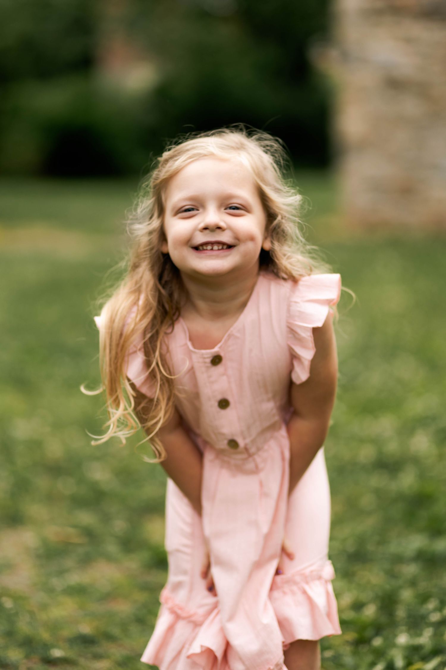 Adorable blonde girl in a pink ruffled dress laughing joyfully on a green lawn outdoors at Lockridge Park.