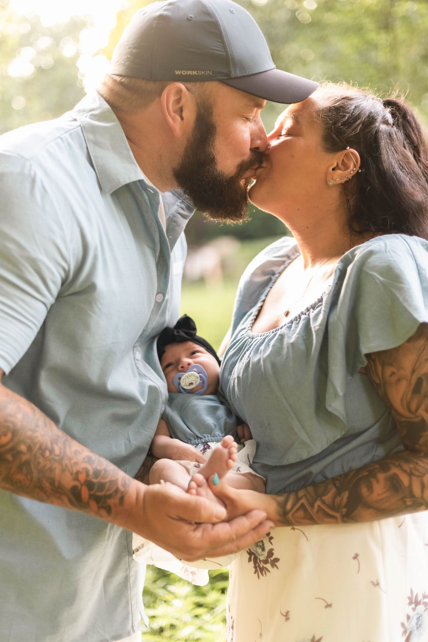 Tattooed couple sharing a kiss while holding their newborn baby with a pacifier in sunny park during a newborn session in Boyertown, PA.