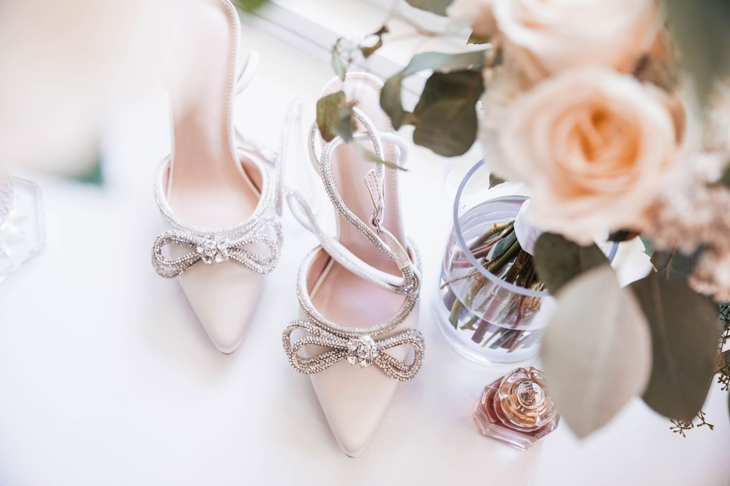 Elegant bridal heels with crystal bow detail beside pink roses and perfume bottle.