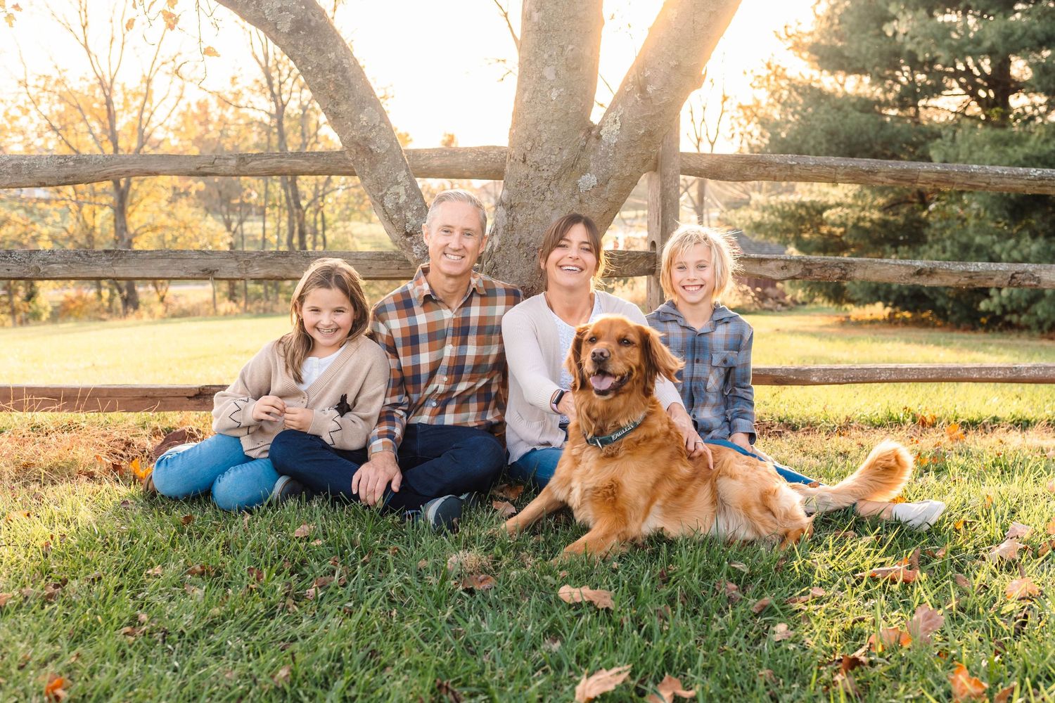 Happy family of four with a golden retriever dog sitting on grass near a wooden fence in fall in Harleysville, PA.