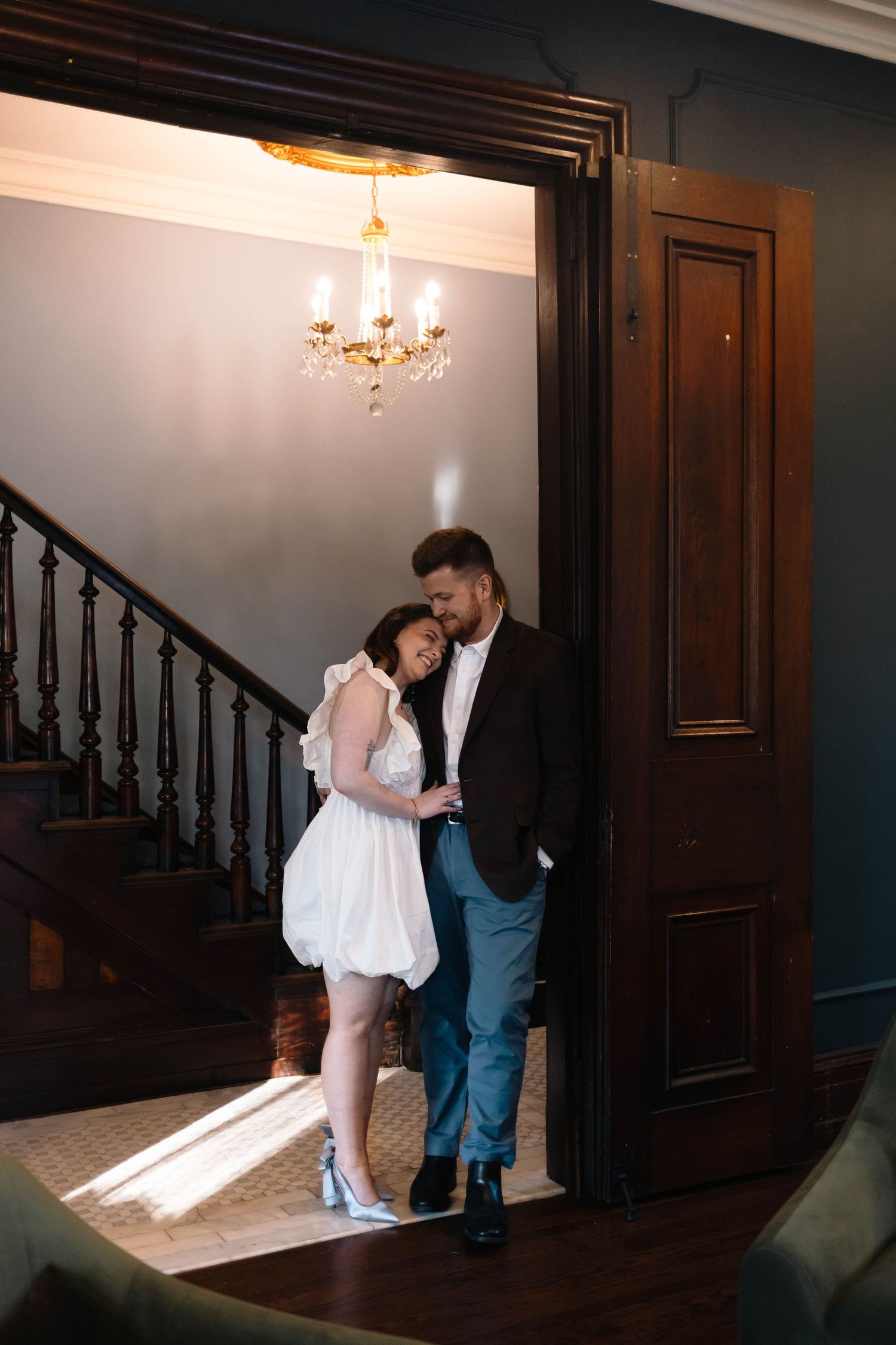 Couple embraces in a sunlit doorway of an elegant home with a crystal chandelier above in West Chester, PA.