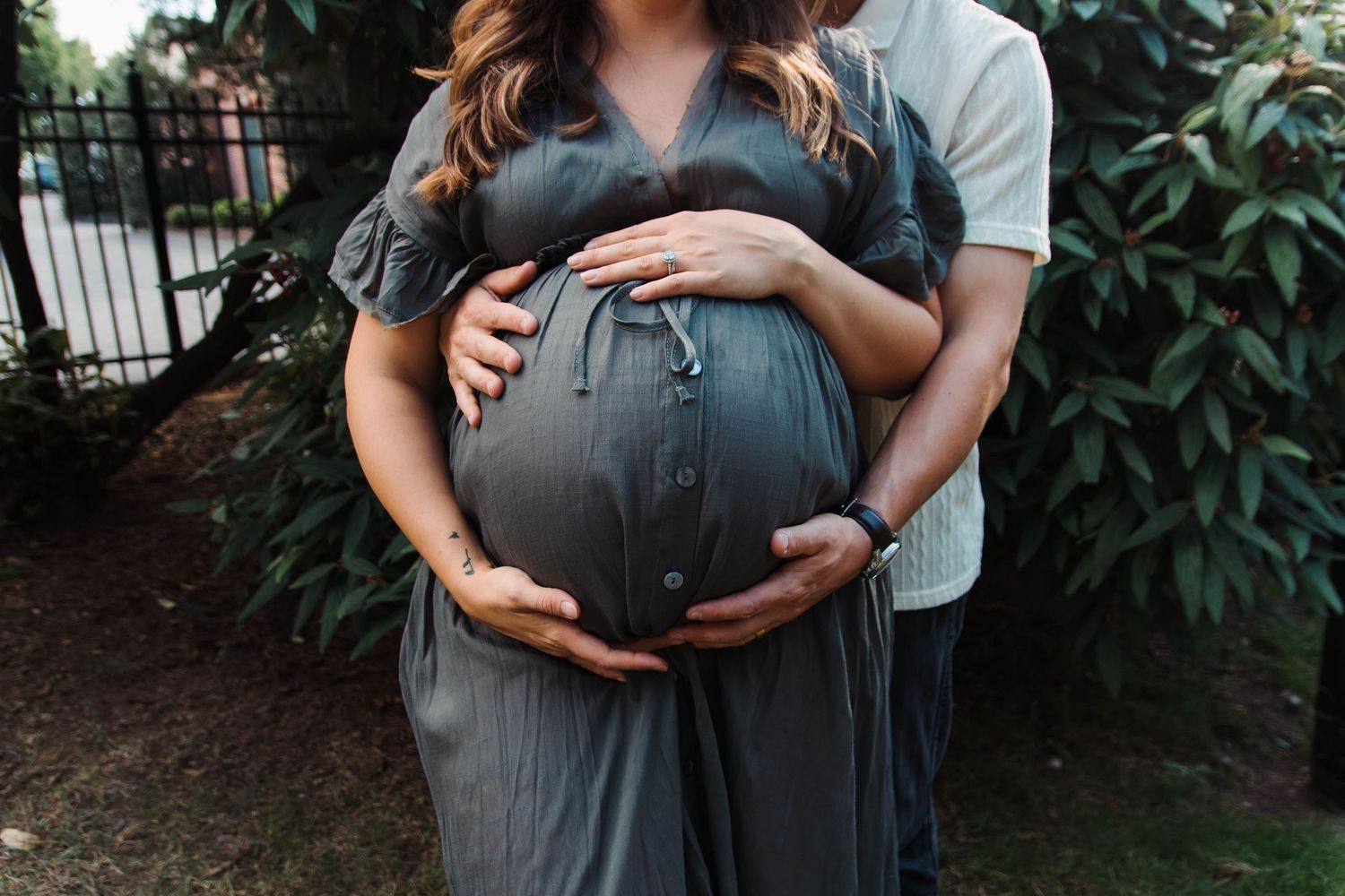 Couple cradling pregnant belly outdoors, wearing a gray dress surrounded by lush greenery.