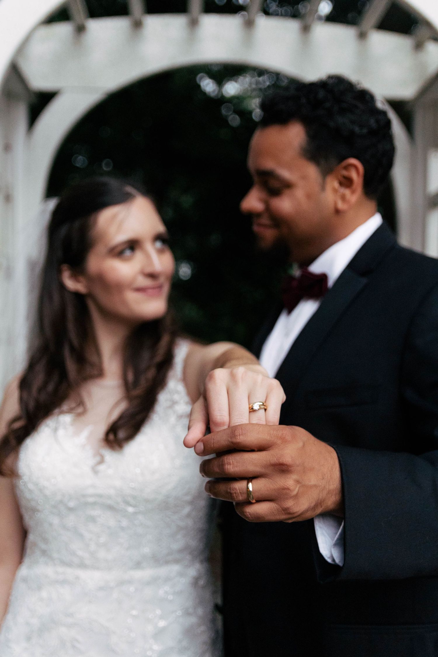 Newlyweds display their wedding rings while holding hands in front of a white garden arbor.