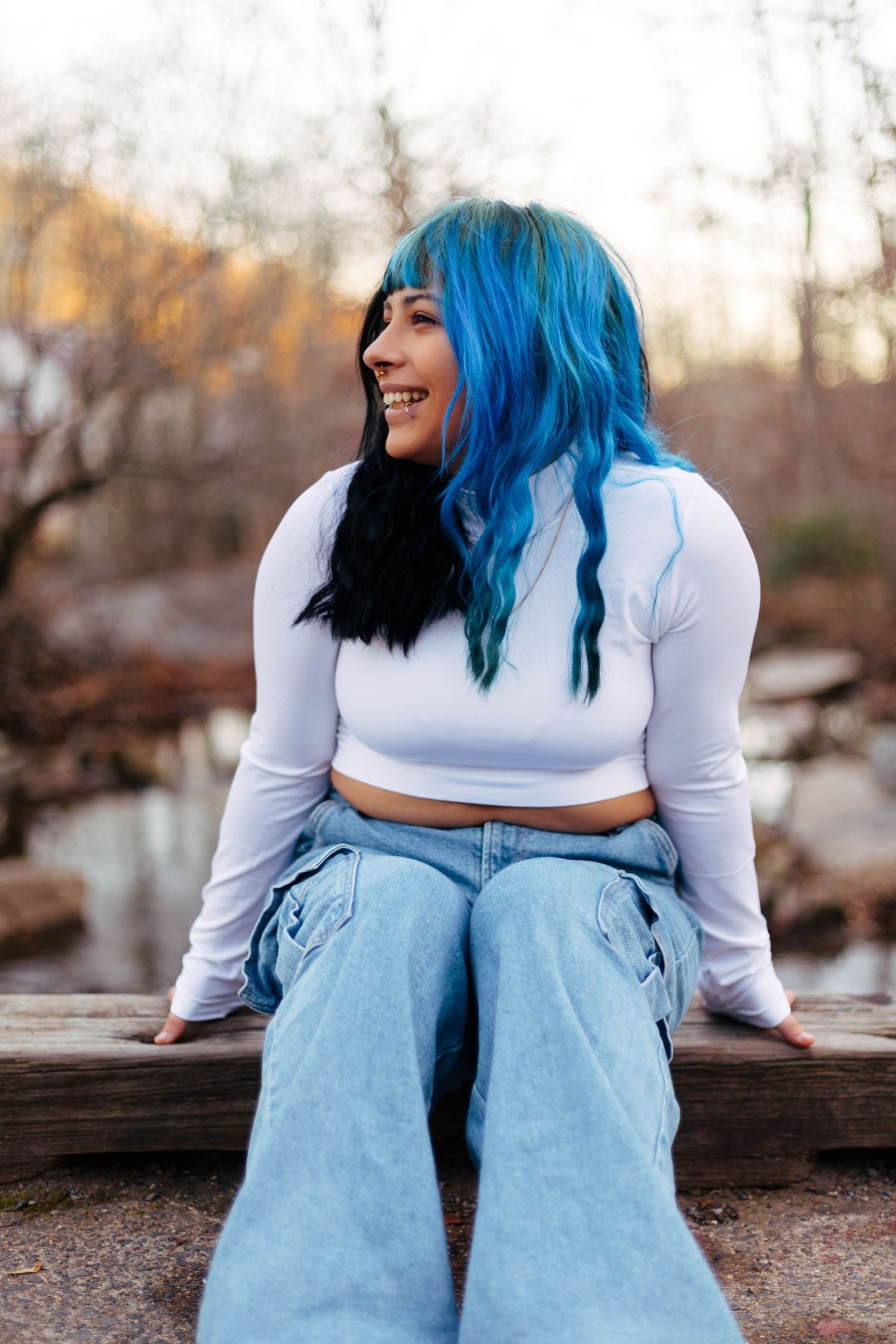 Smiling young woman with blue and black hair wearing white crop top and wide-leg jeans outdoors.
