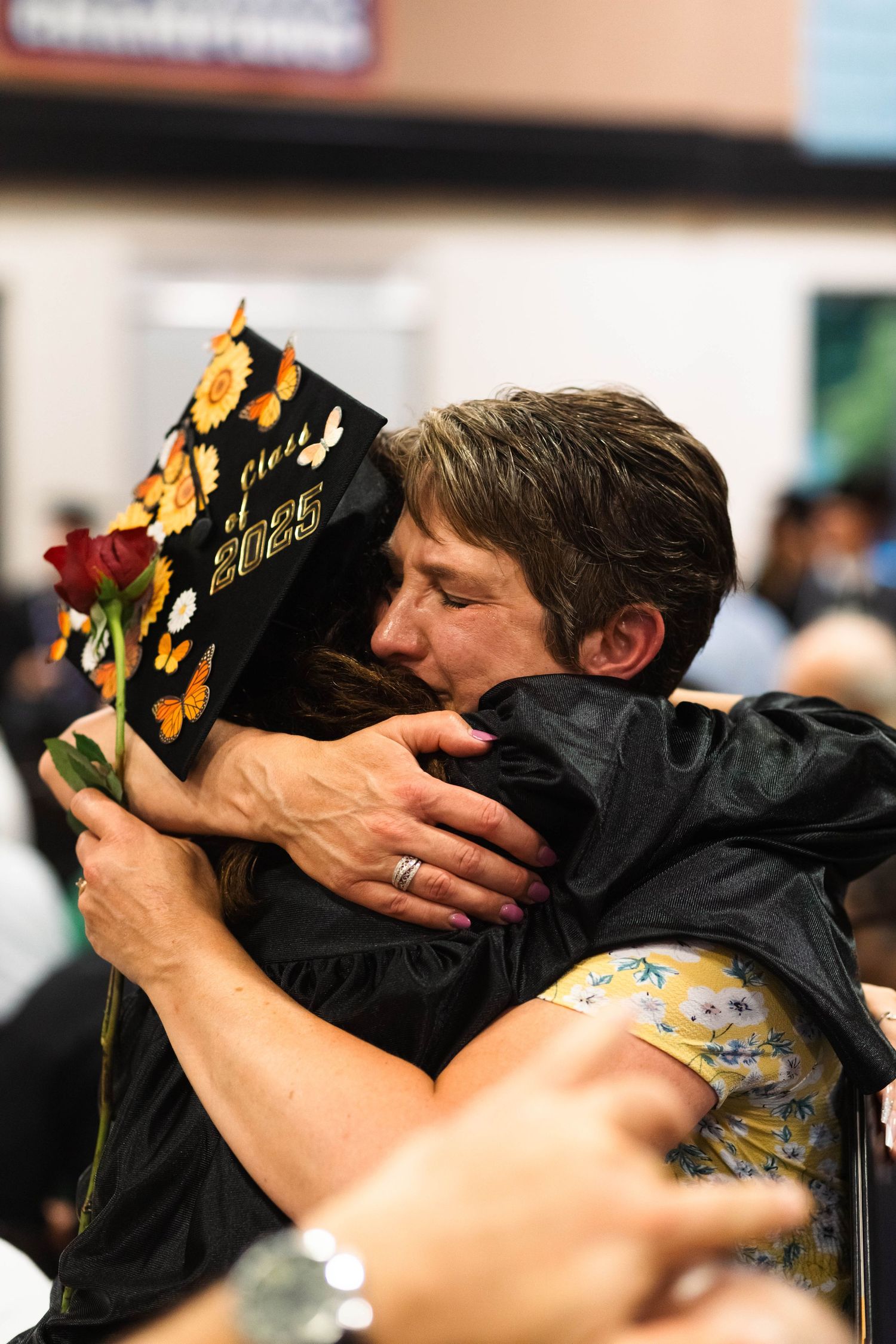 Graduate in cap and gown hugging loved one while holding red rose at ceremony.