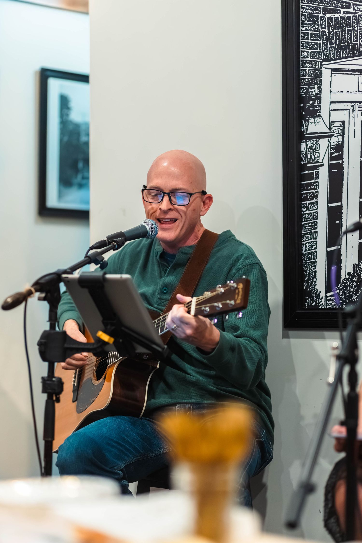 Bald man with glasses playing acoustic guitar and singing into a microphone at an indoor performance.