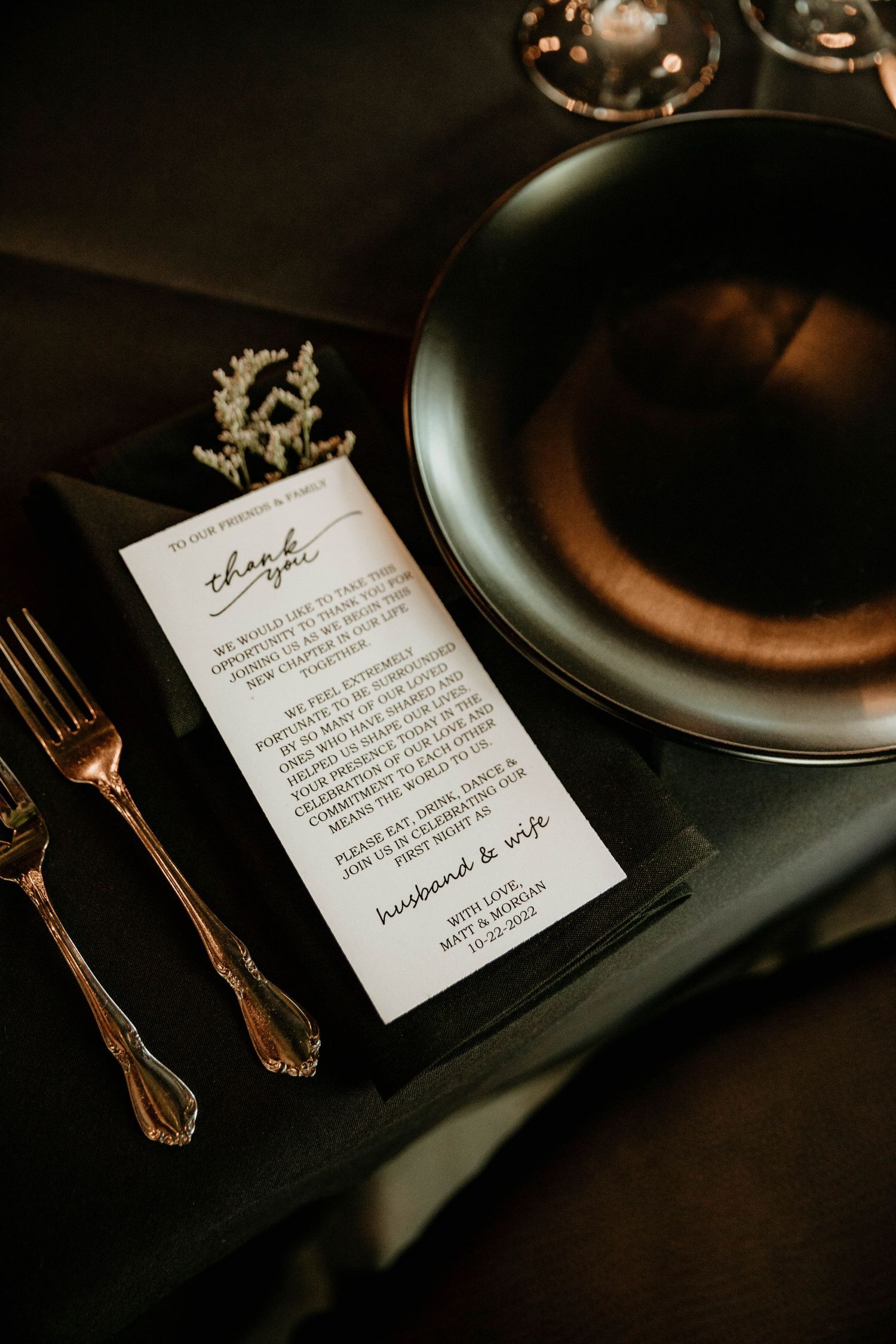 Elegant dark wedding place setting with gold cutlery, black plate, and thank you card.
