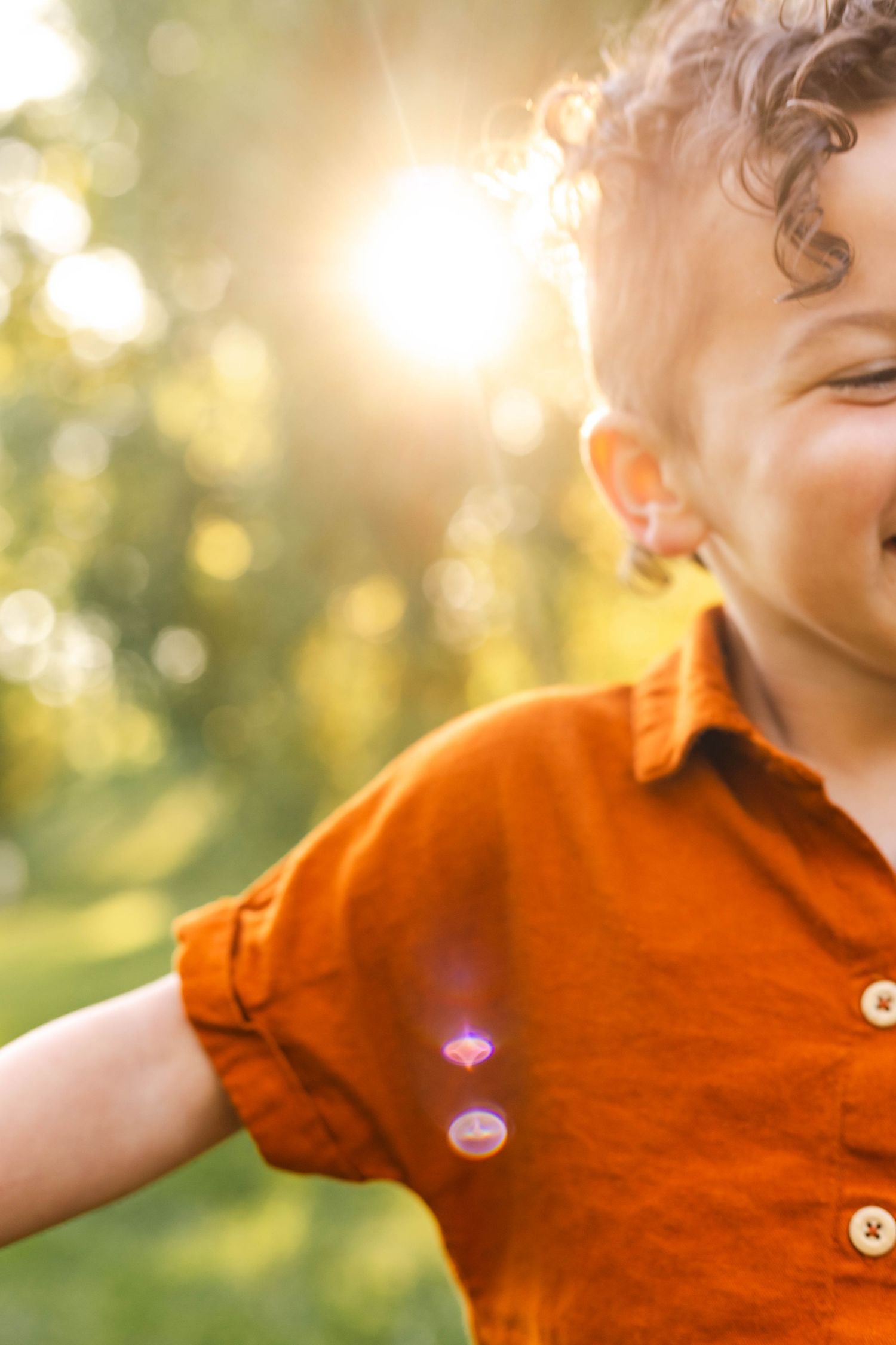 Happy toddler in orange shirt laughing outdoors in warm golden sunlight.