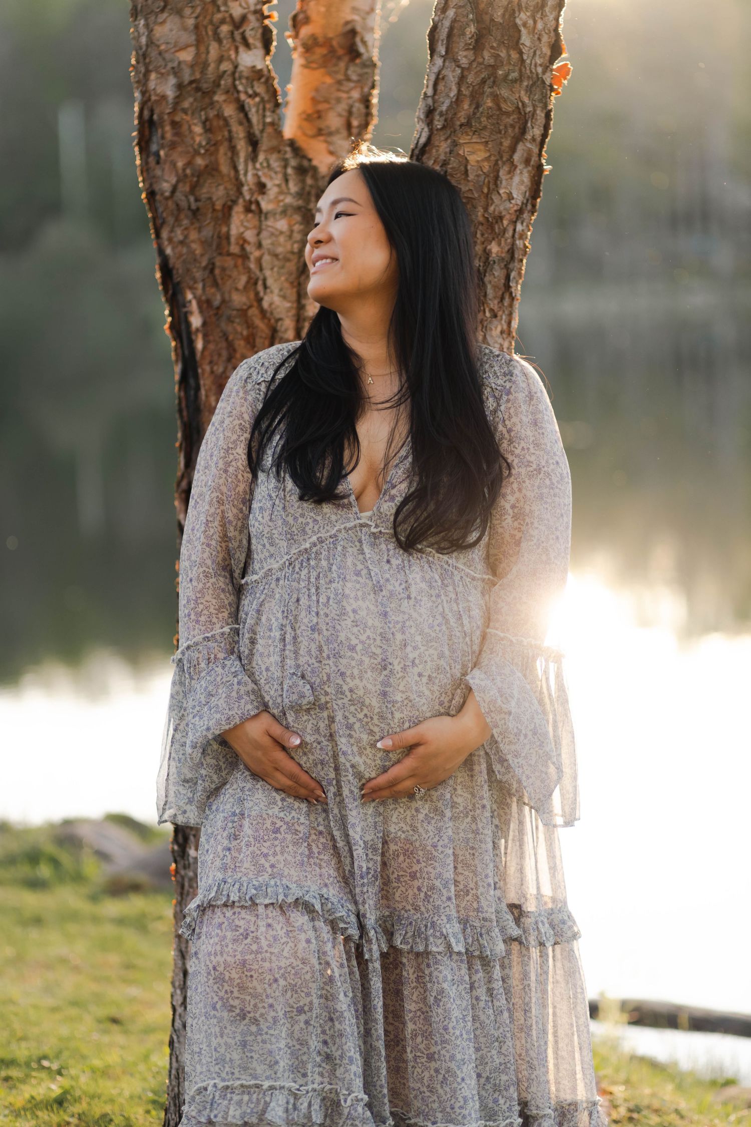 Smiling pregnant woman in floral dress cradling her baby bump while standing by a sunlit tree near a lake.