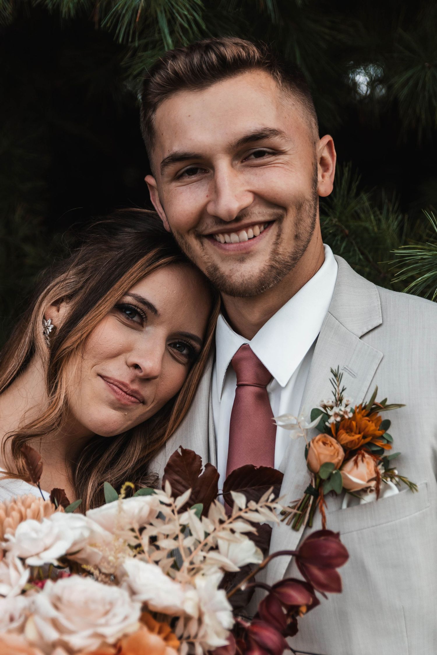 Smiling bride and groom pose closely together with a lush floral bouquet and pine trees behind them.