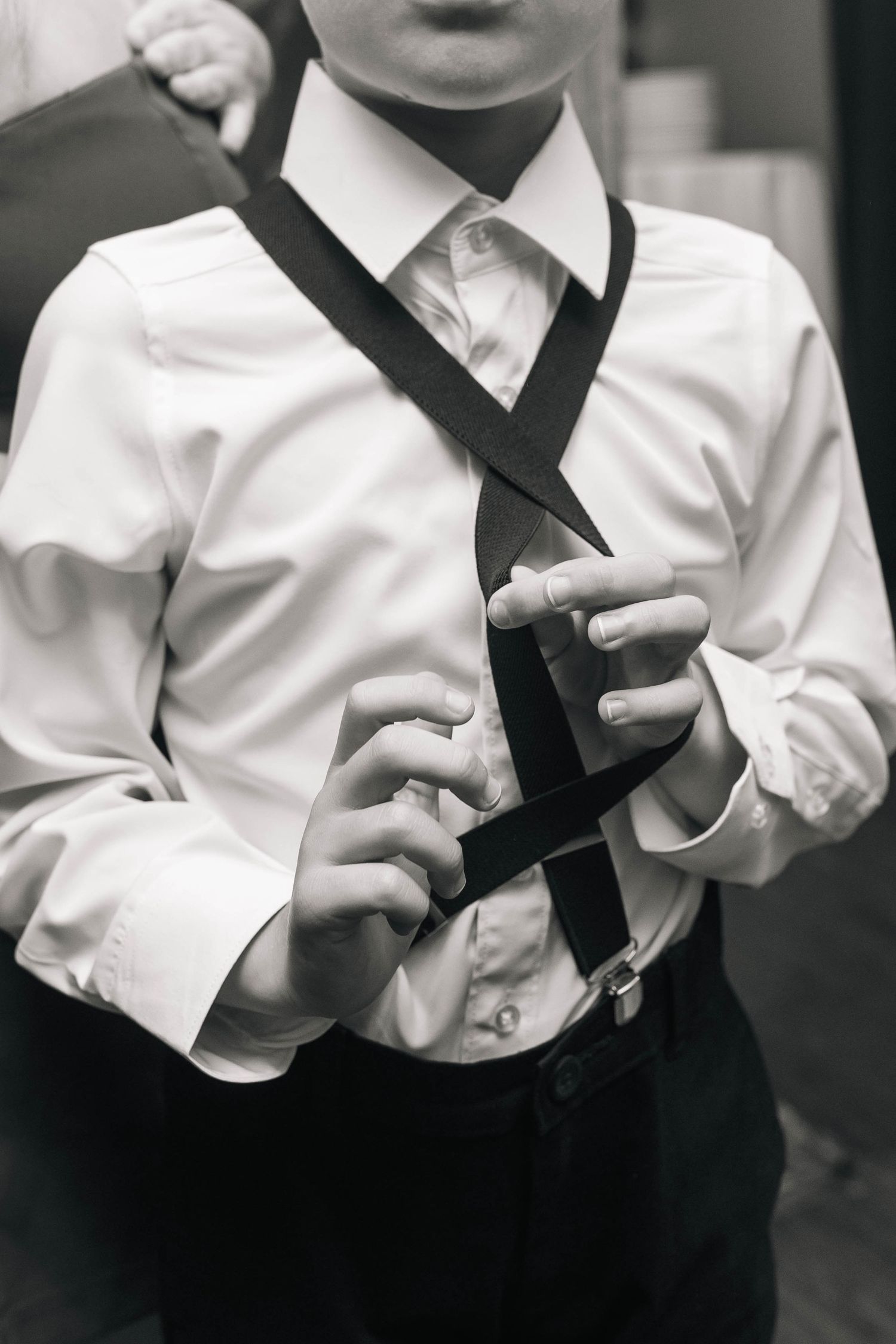 Black and white photo of young ring bearer in white dress shirt adjusting suspenders at wedding.