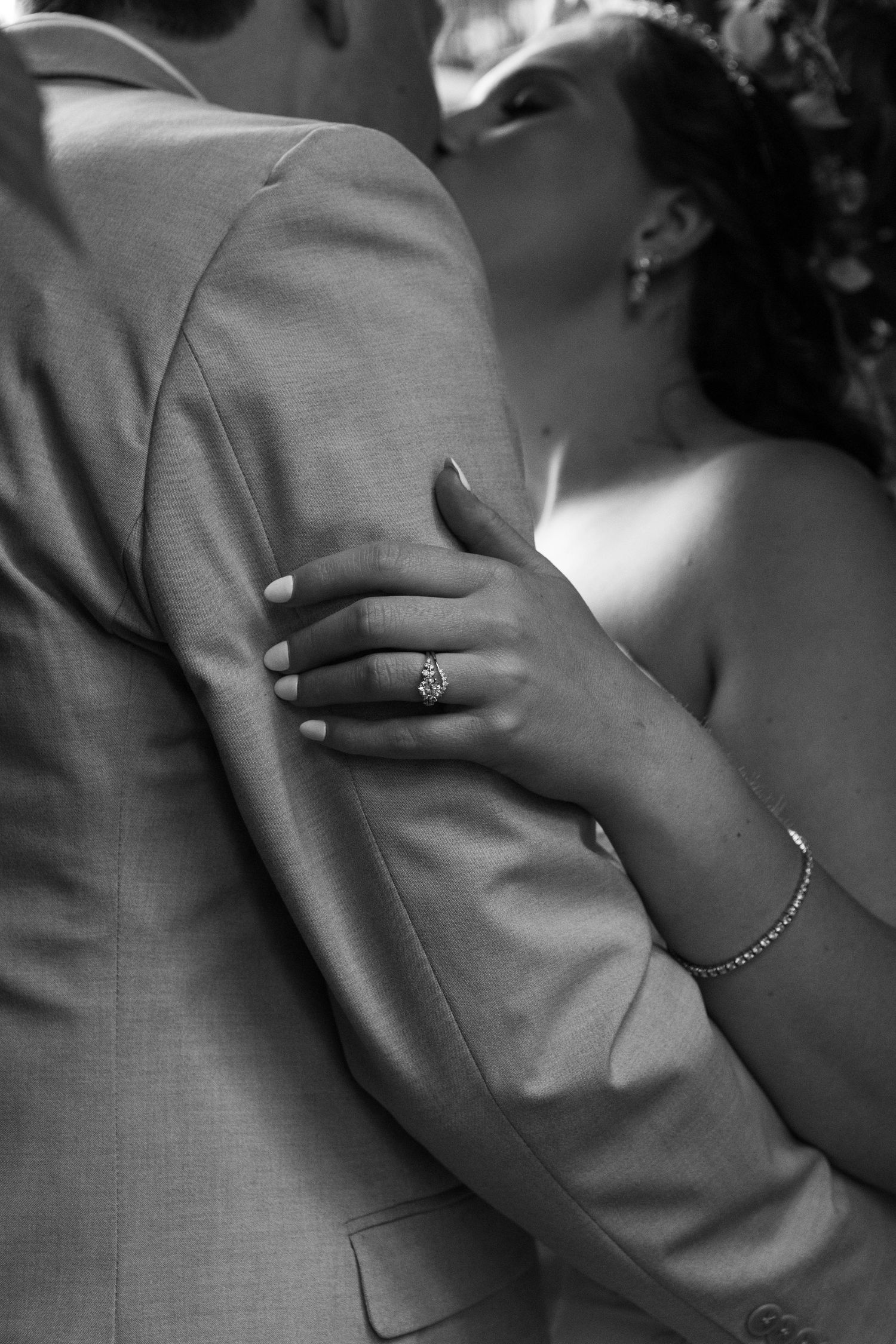 Black and white close-up of bride's hand with diamond ring and bracelet resting on groom's tan suit jacket.