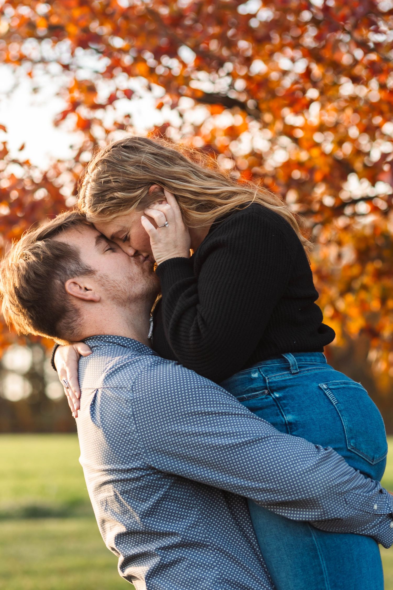 Man lifting and kissing woman in front of vibrant red autumn tree during fall engagement session in Lancaster, PA.