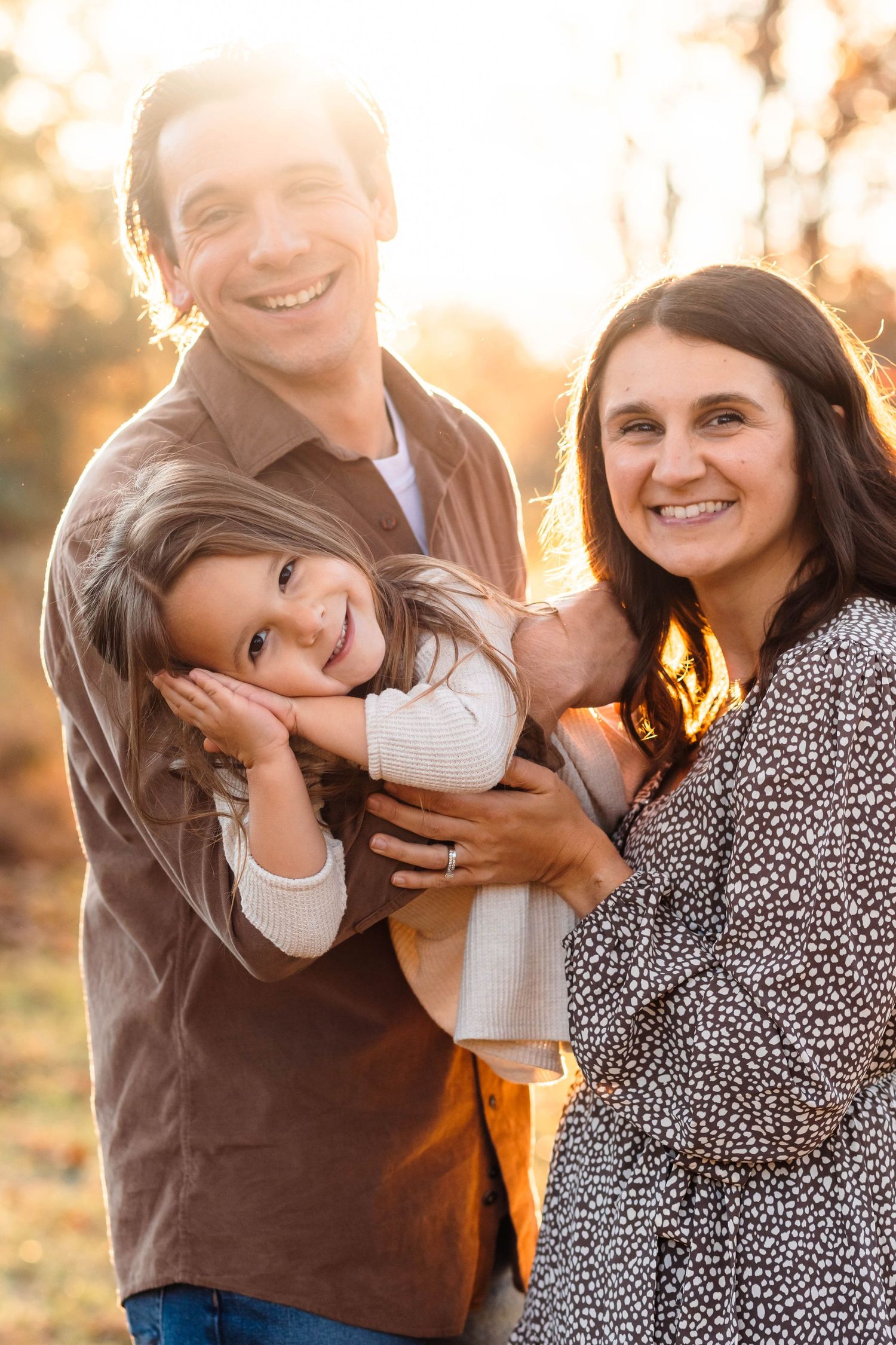 Smiling family of three poses outdoors during golden hour, parents holding young child in warm autumn light in Pottstown, PA.
