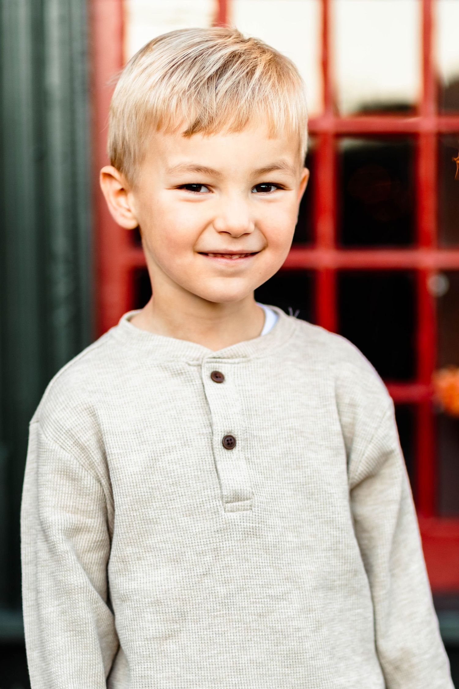 A young blonde boy in a gray henley shirt smiles softly in front of a red-framed window at the Colebrookdale Railroad Station in Boyertown.