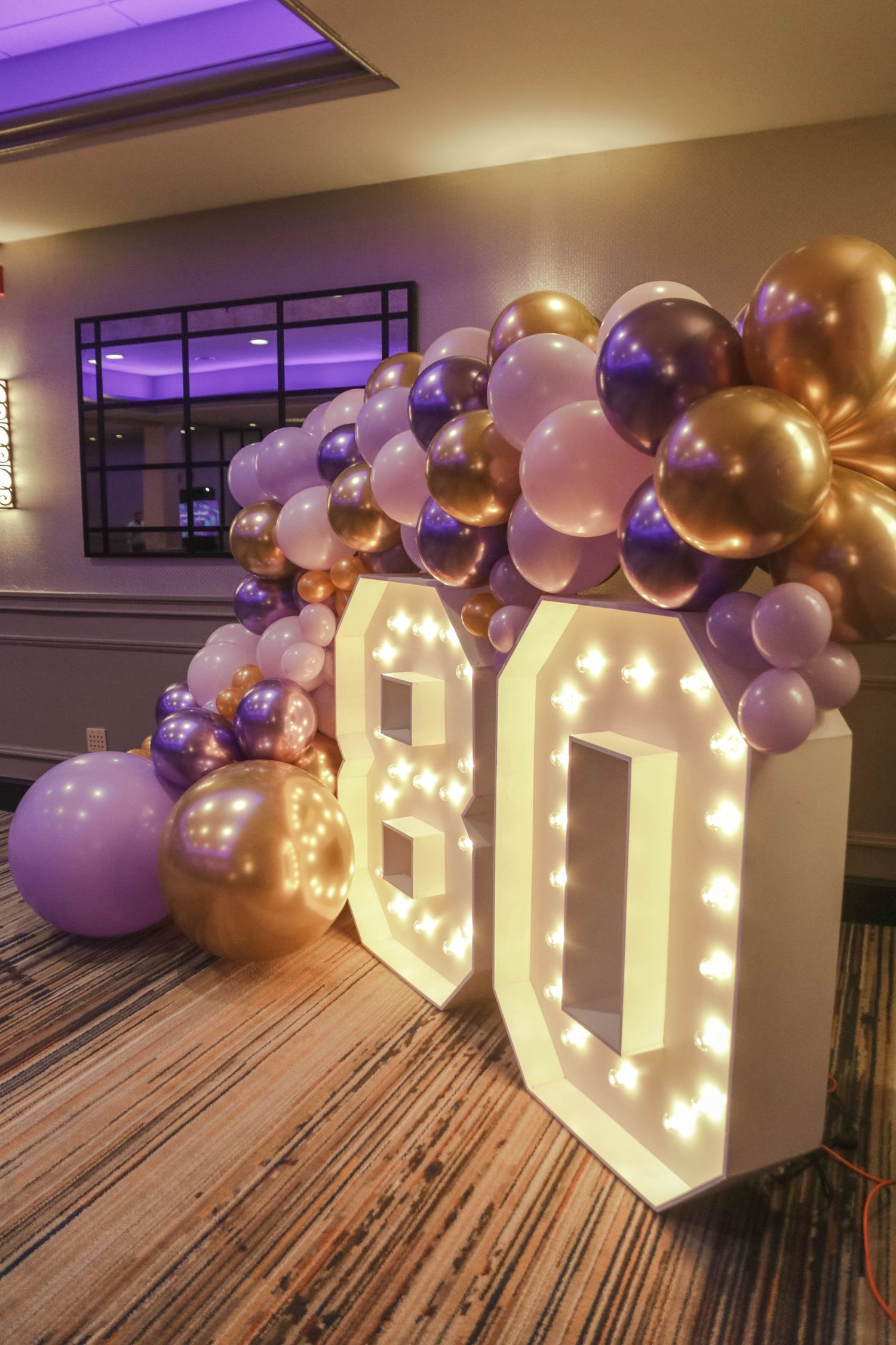 Illuminated marquee number 80 surrounded by purple, lavender, and gold balloon garland for a birthday party.