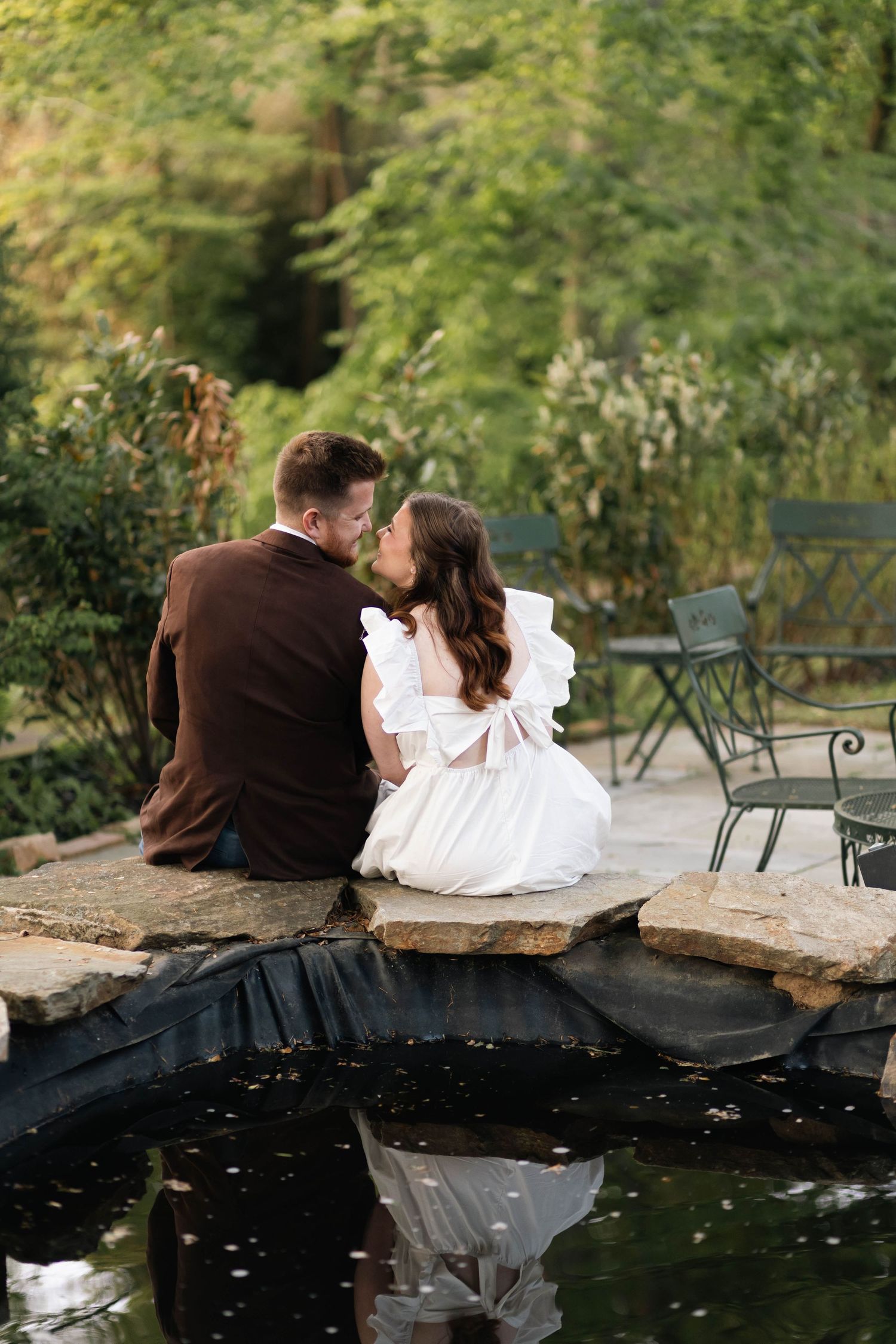 Engaged couple sitting close together on stone bridge over a reflective garden pond in West Chester, PA.