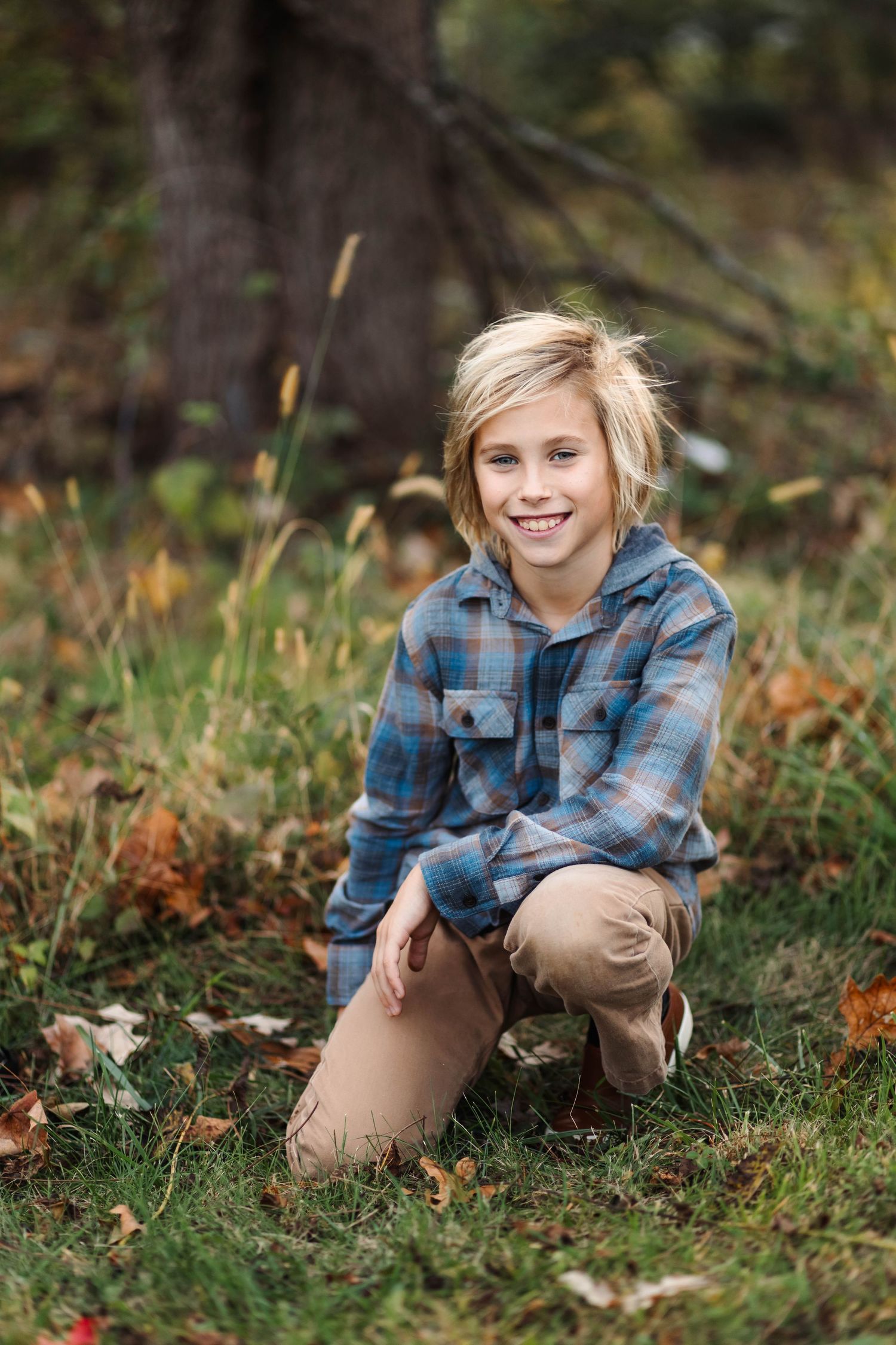 A blonde boy crouches on grass surrounded by fallen autumn leaves and smiles at the camera in Harleysville, PA.