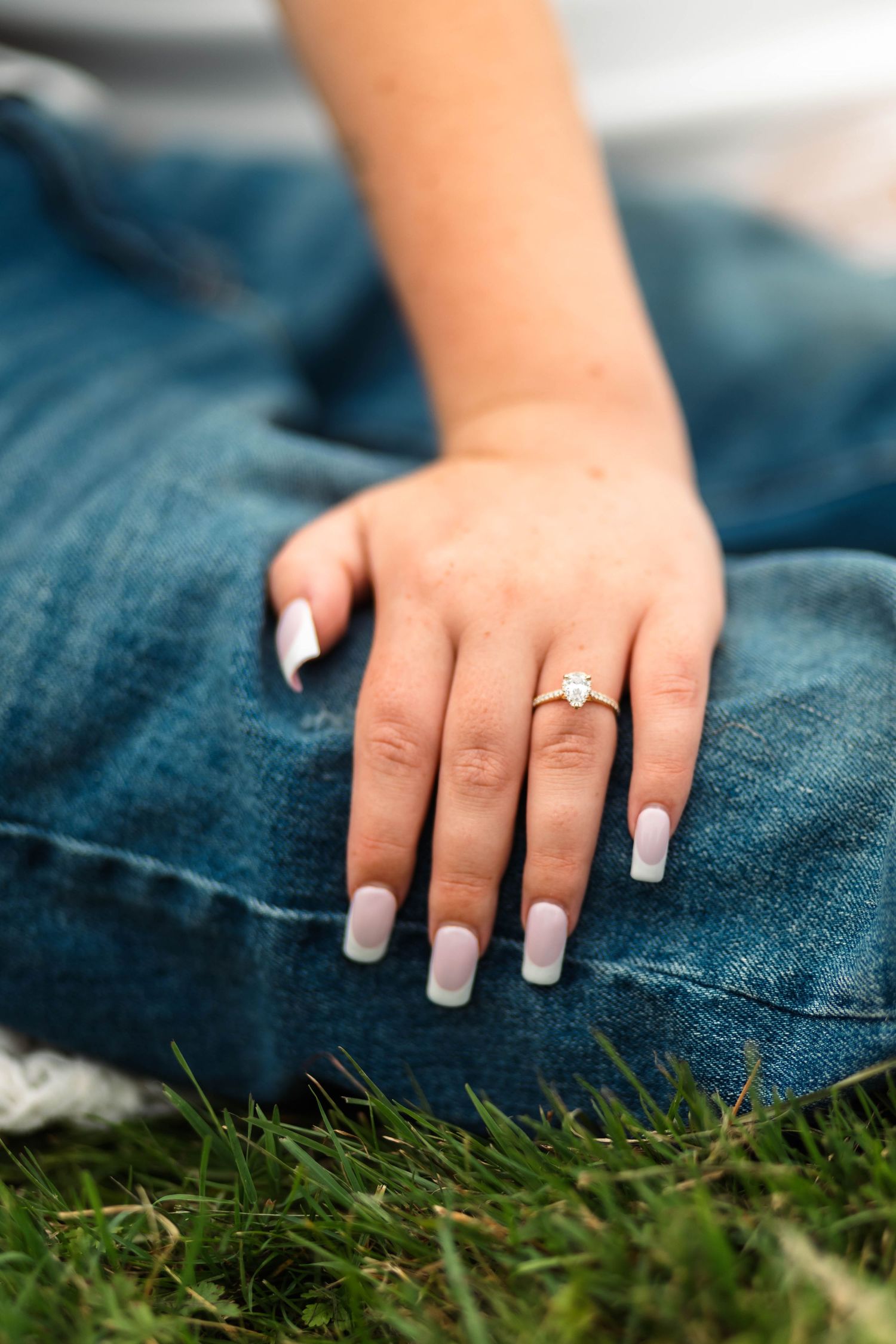 Close-up of a woman's hand with French manicure nails and diamond engagement ring resting on blue denim jeans during an engagement session.