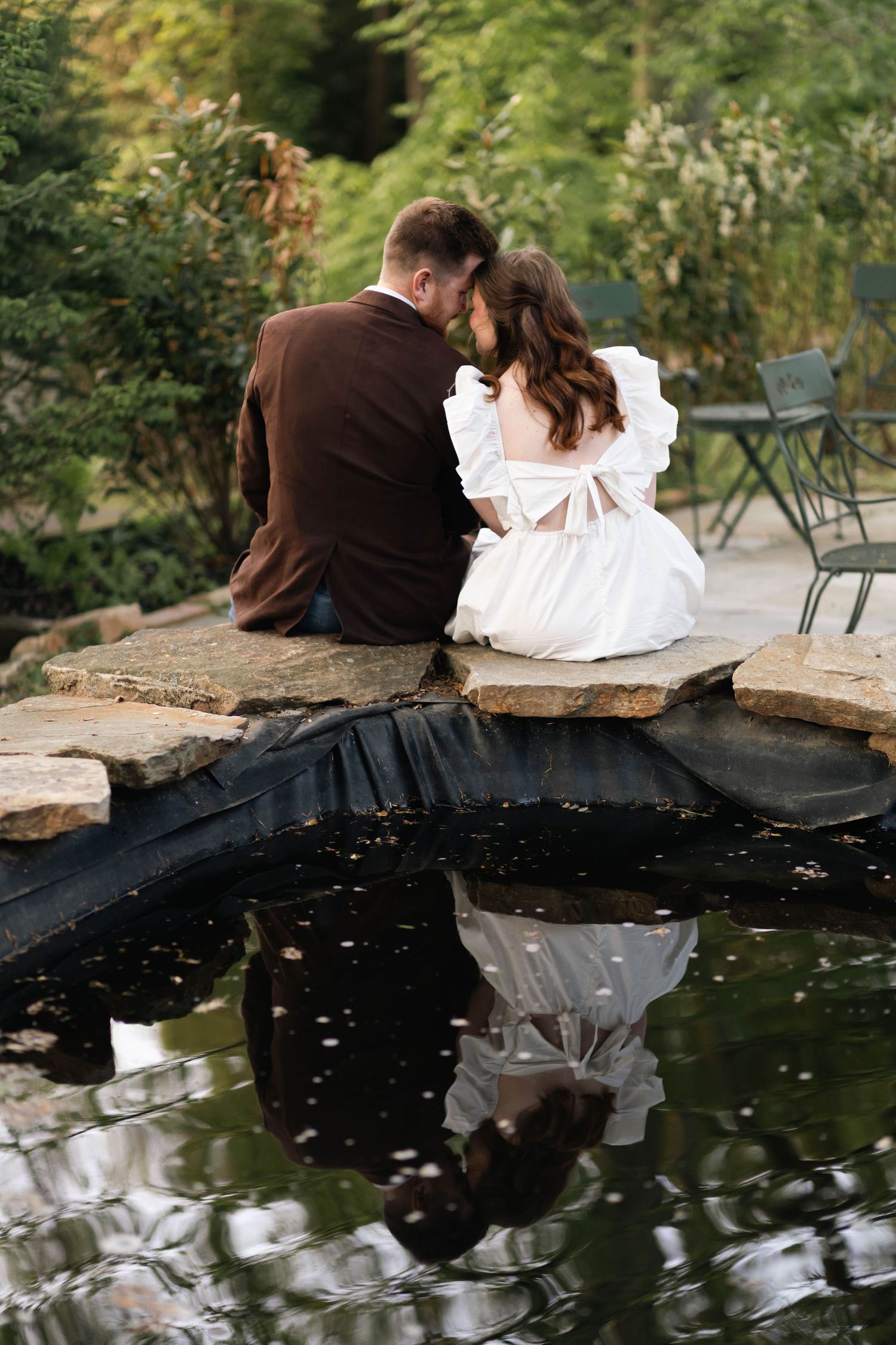 Bride and groom share intimate moment seated by reflective garden pond, their reflection visible in still water in West Chester, PA.
