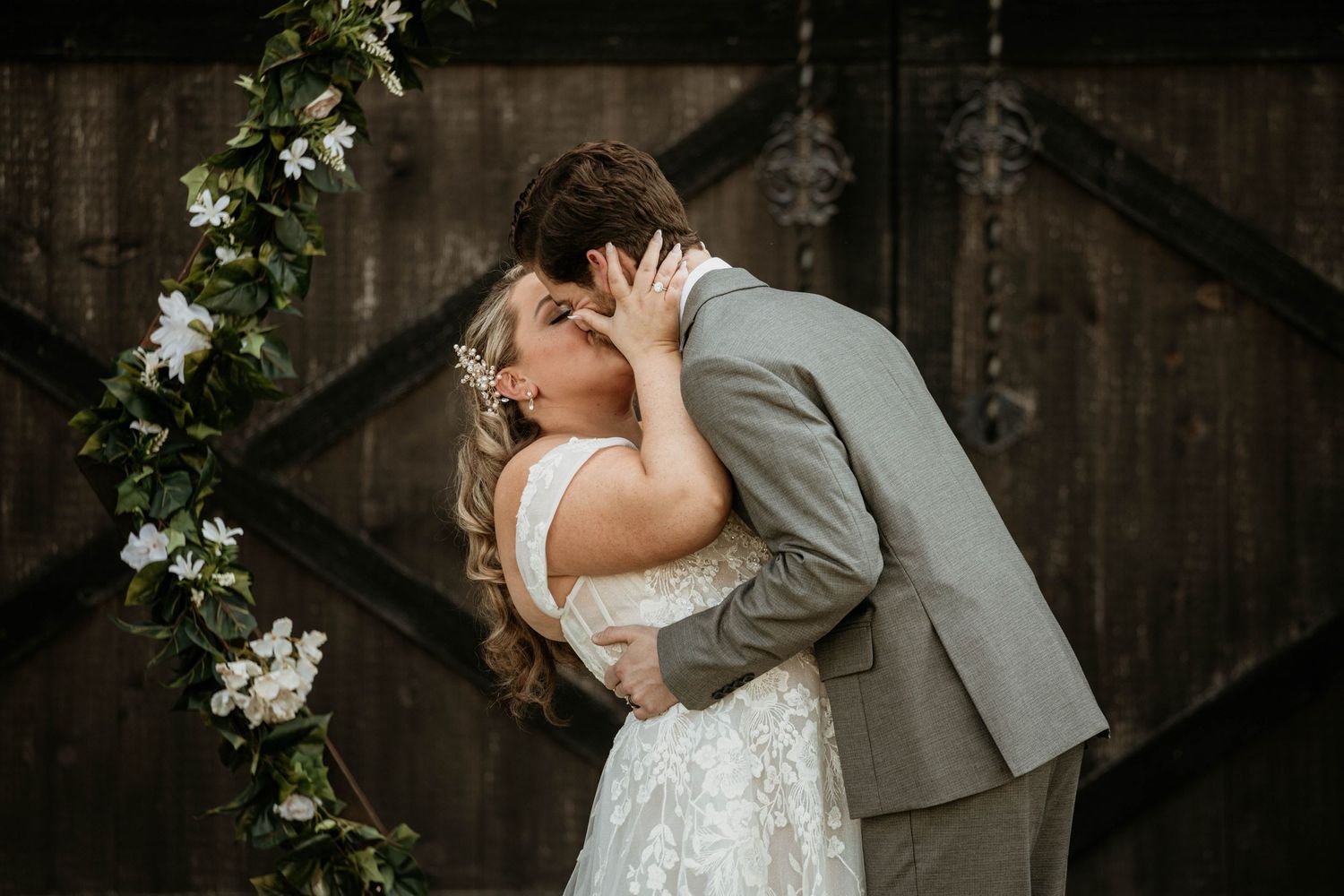 Bride and groom kissing in front of floral arch against rustic wooden barn door.