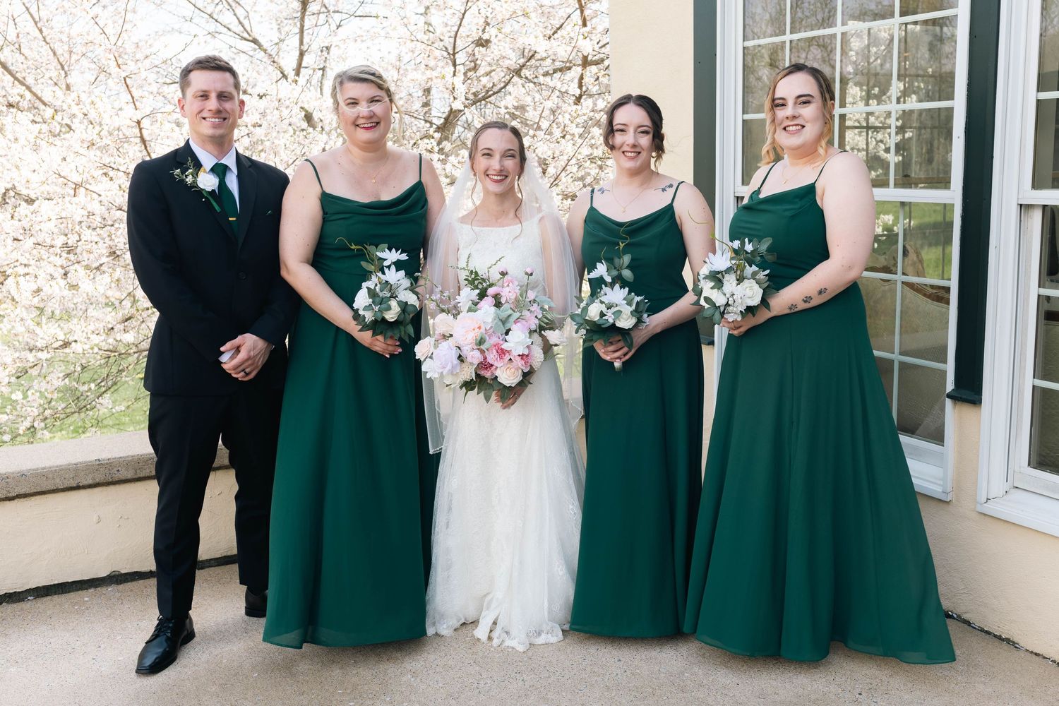 Bride with bridesmaids in emerald green gowns and groomsman pose outdoors near spring blossoms.