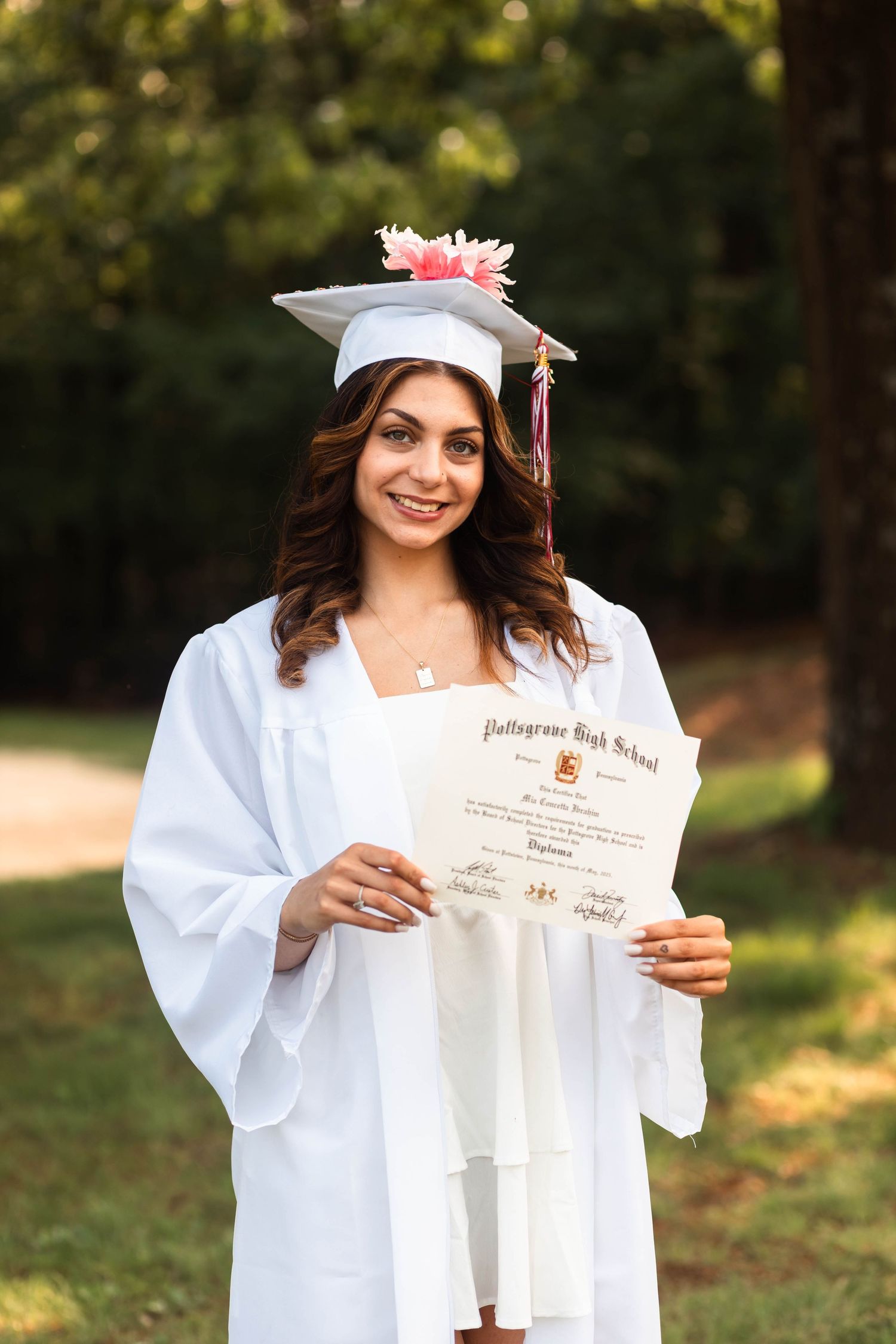 Pottsgrove high school graduate in white cap and gown holding her Pottsgrove High School diploma outdoors in nature.