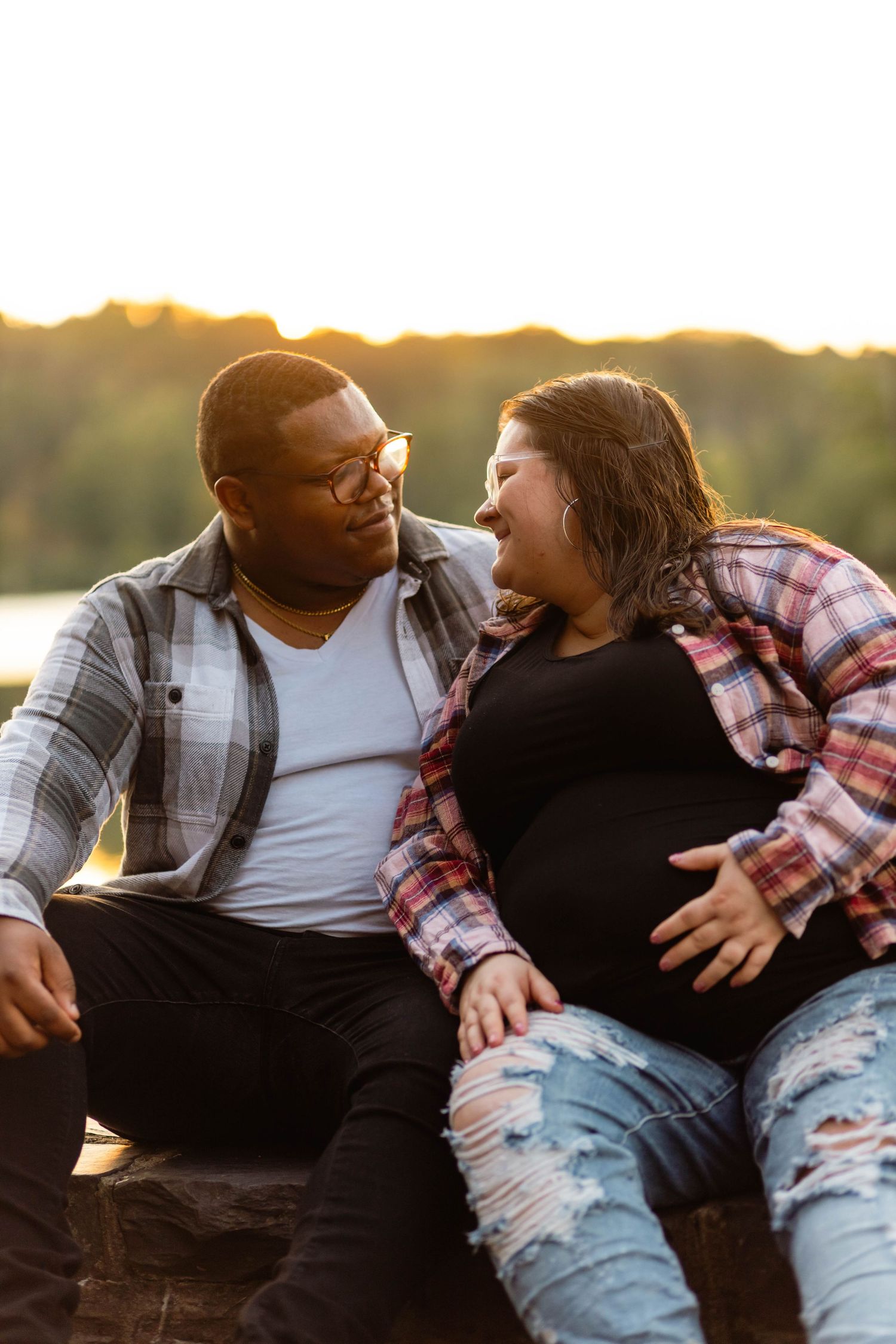 Happy interracial couple sitting together at sunset, woman showing pregnant belly in warm golden light at Green Lane Park.