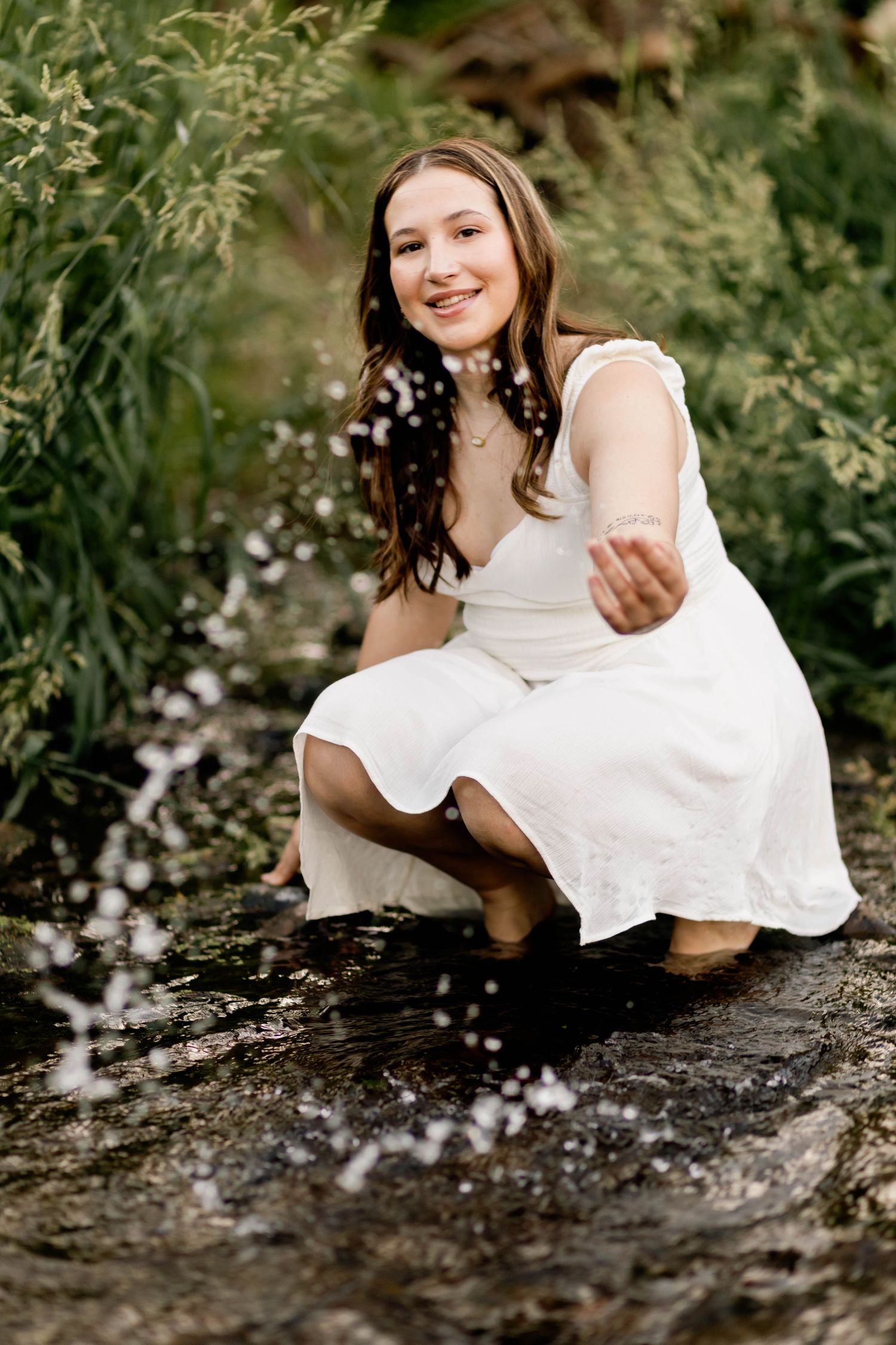 Boyertown Senior in white dress splashes water while crouching in a stream in Harleysville, PA.