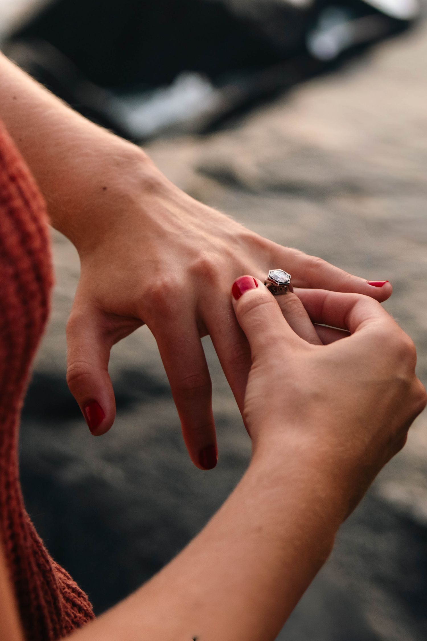 Close-up of a man placing a diamond engagement ring on a womans finger with red nail polish at the beach.