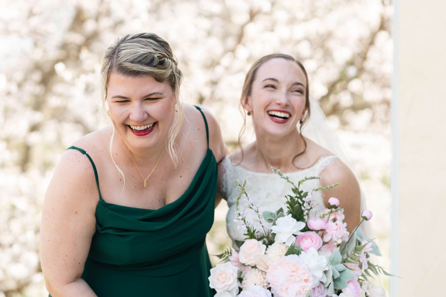 Bride and bridesmaid in green dress share a joyful laugh together outdoors at spring wedding.