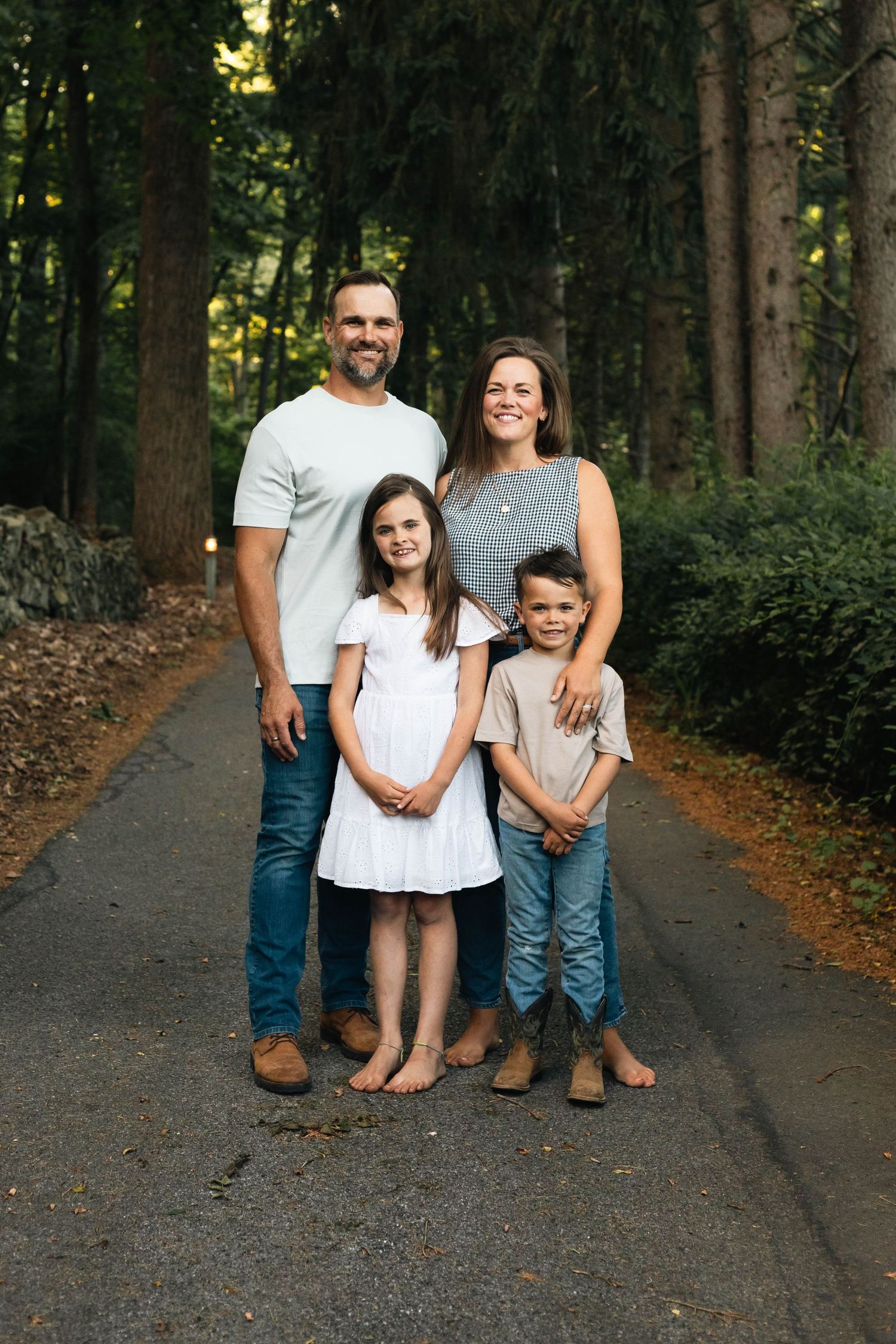 Happy family of four posing barefoot on a tree-lined path, parents with young daughter and son smiling.