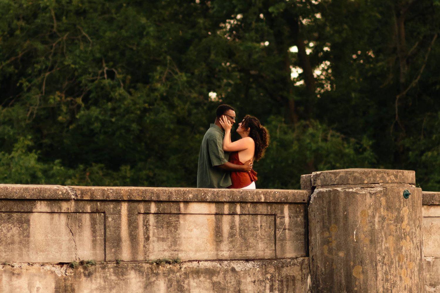 Romantic couple kissing on a stone wall bridge surrounded by lush green trees at dusk at Grings Mill in Reading, PA.